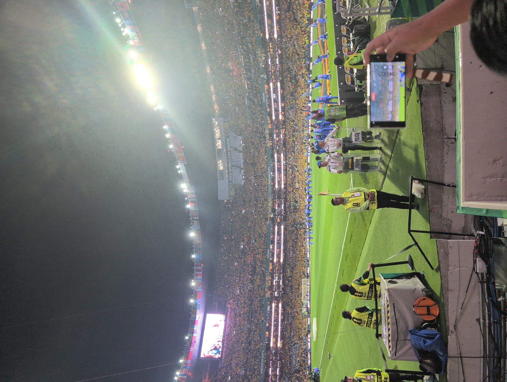 A panoramic view of a large stadium at night shows a soccer match. The stadium is filled with spectators dressed primarily in yellow. In the foreground, several security guards in yellow vests are standing on the sidelines. A large screen displays the words "MEXICO CITY" and "FIFA WORLD CUP 2026". The field is illuminated by bright stadium lights. In the bottom right corner, a hand is holding a smartphone, recording the event. The overall scene depicts a vibrant and energetic atmosphere at a major sporting event.