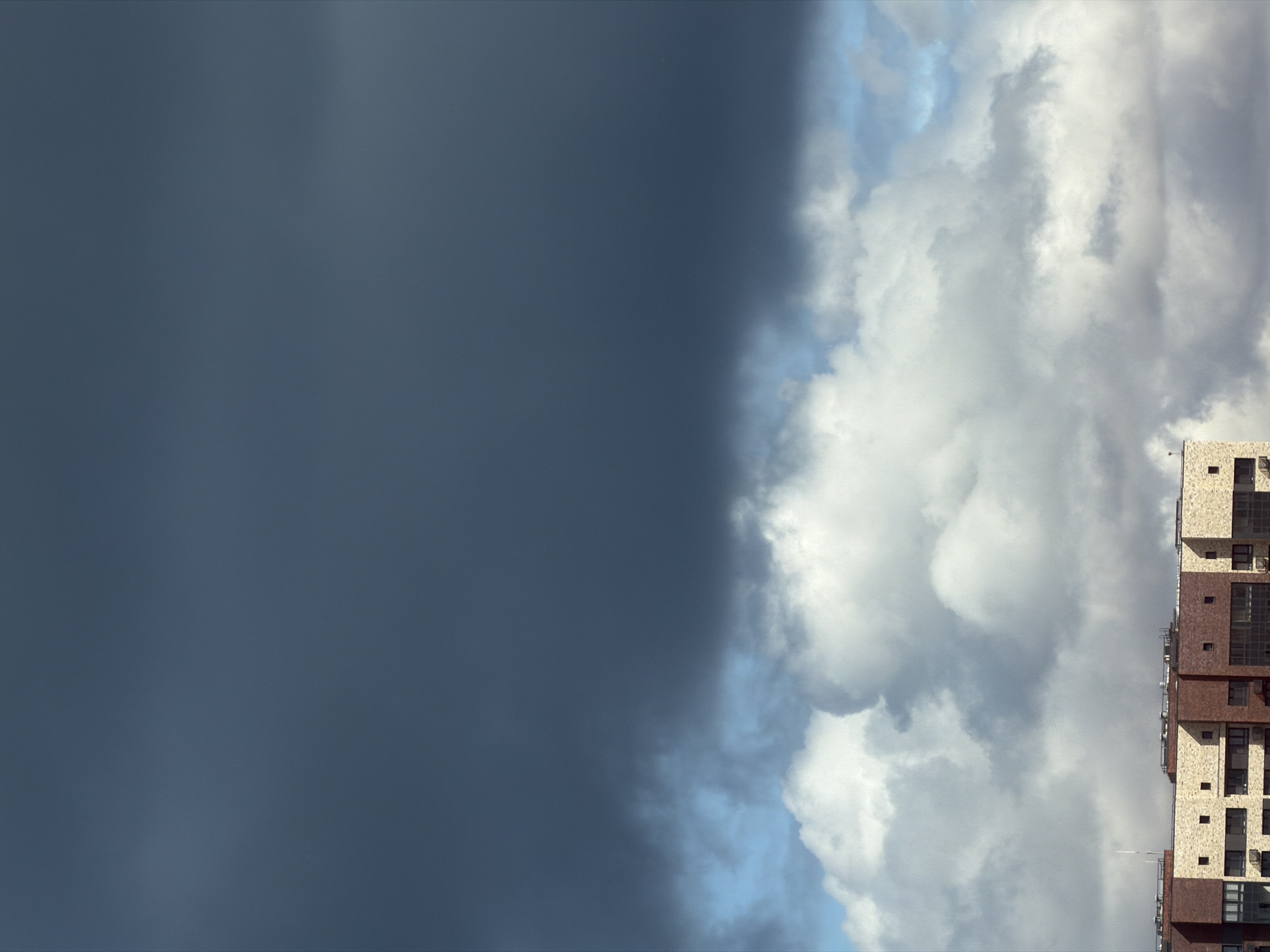 The image shows a view of a dark, stormy sky on the left side and a bright, cloudy sky on the right side, separated by a distinct line. A multi-story building is visible on the right edge of the image. The location is the city of Moscow, Russia.