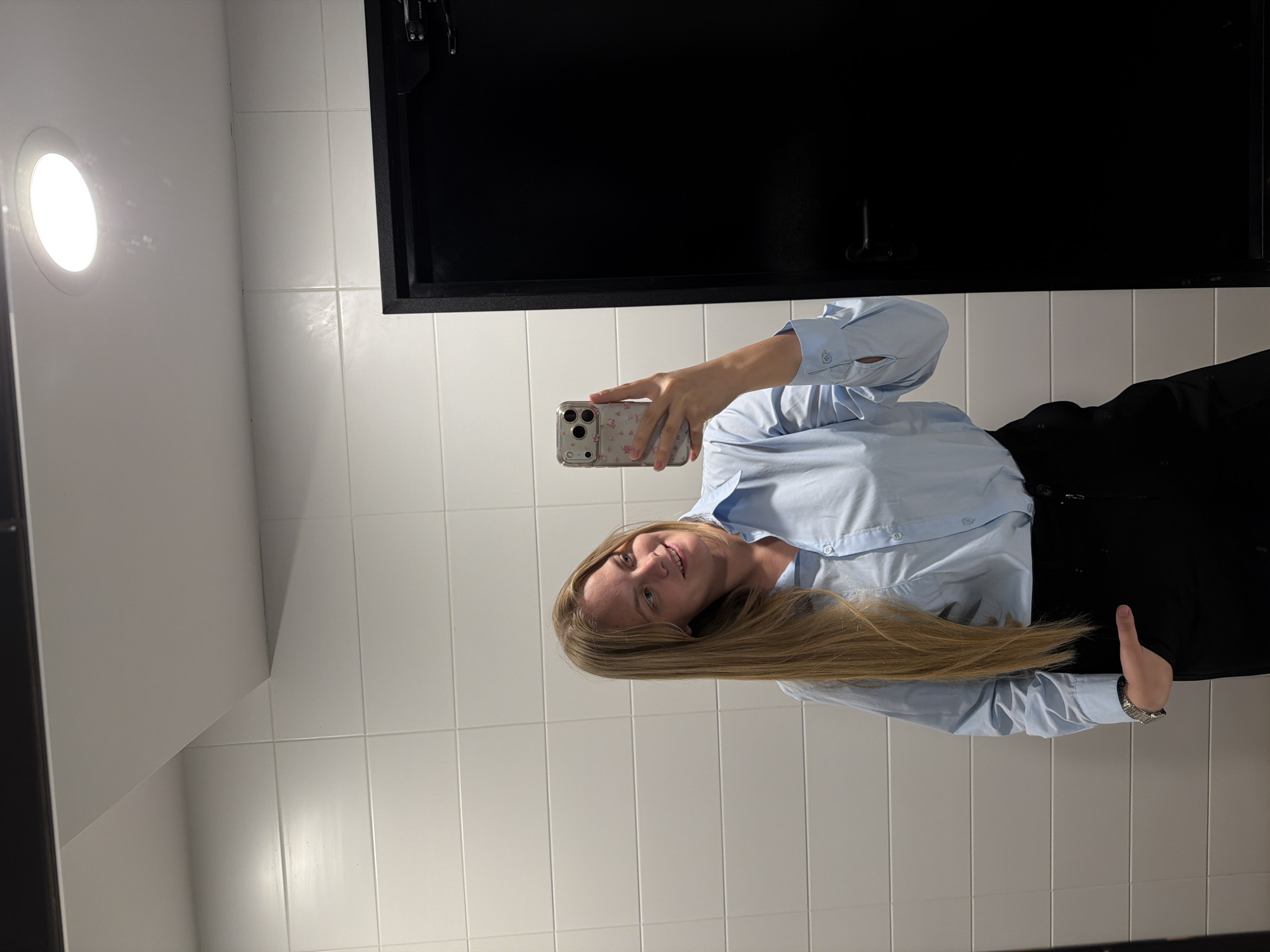 A person is taking a selfie in a bathroom. The bathroom has white tiled walls and a black framed mirror or cabinet. The person is wearing a light blue button-down shirt and black pants. They are holding a smartphone with a floral patterned case in their right hand to take the photograph. The person appears to be Caucasian with long blonde hair. A round light fixture is visible on the wall. The location is not identifiable as Izhevsk, Russia.