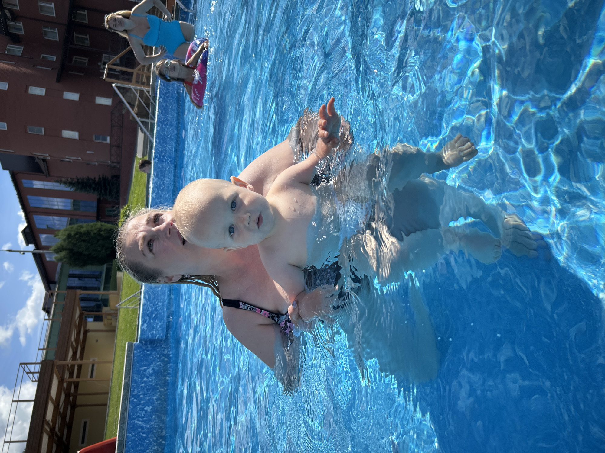 A woman and a toddler are in a swimming pool, with the woman holding the toddler. The toddler is facing the camera and has blue eyes. The woman is looking to the side. Other people are visible in the pool in the background. The pool is blue and clear, with sunlight reflecting off the surface. Buildings and trees are visible in the background. The location appears to be an outdoor swimming pool complex.