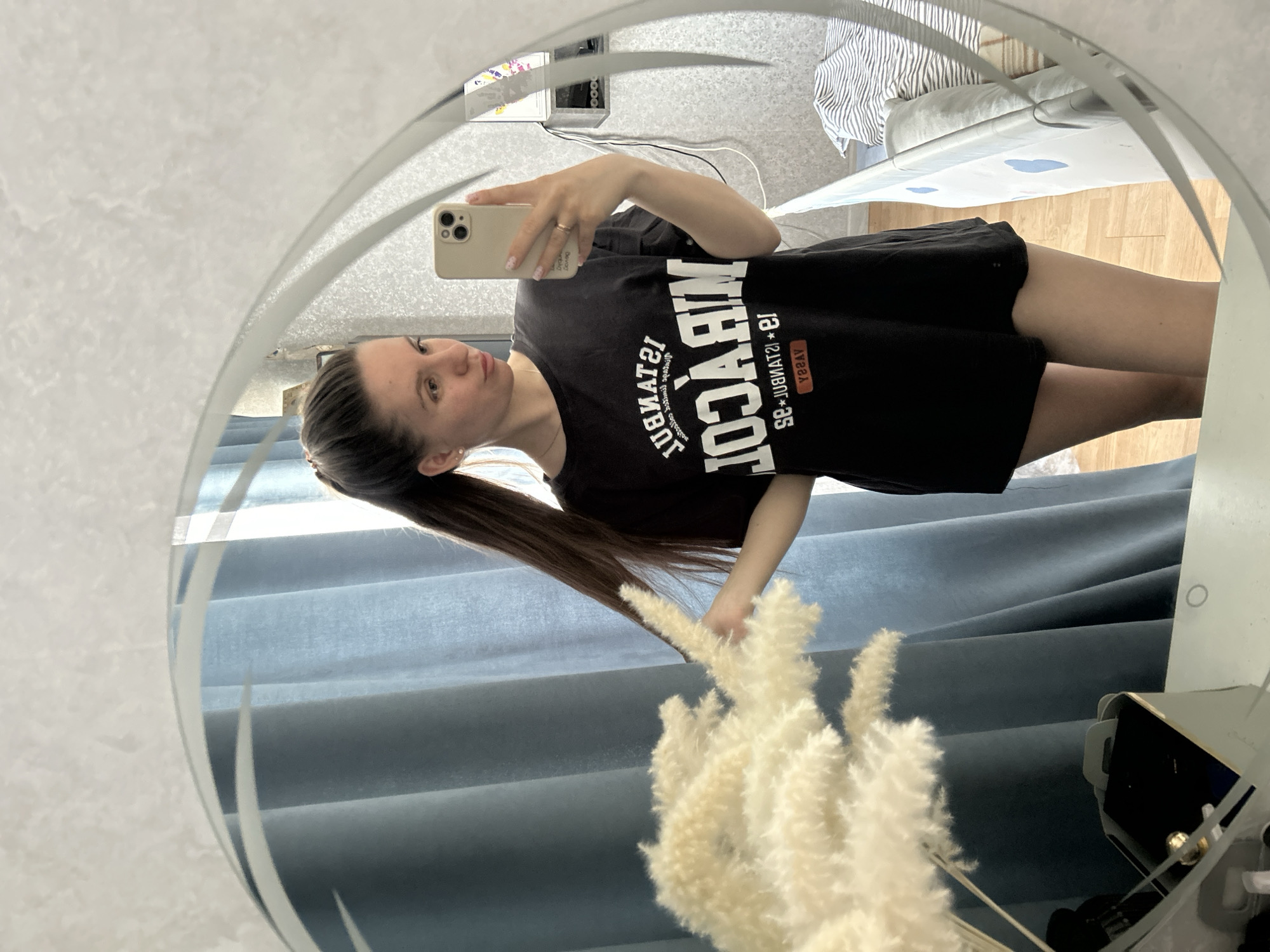 A woman is reflected in a round mirror. She is holding a phone and taking a photo of herself. She is wearing a black t-shirt and dark shorts. Her hair is pulled back into a ponytail. Behind her are blue curtains. In front of her, partially obscuring the view, is a vase filled with white pampas grass. The location is likely not Klin, Russia, based on available visual information.
