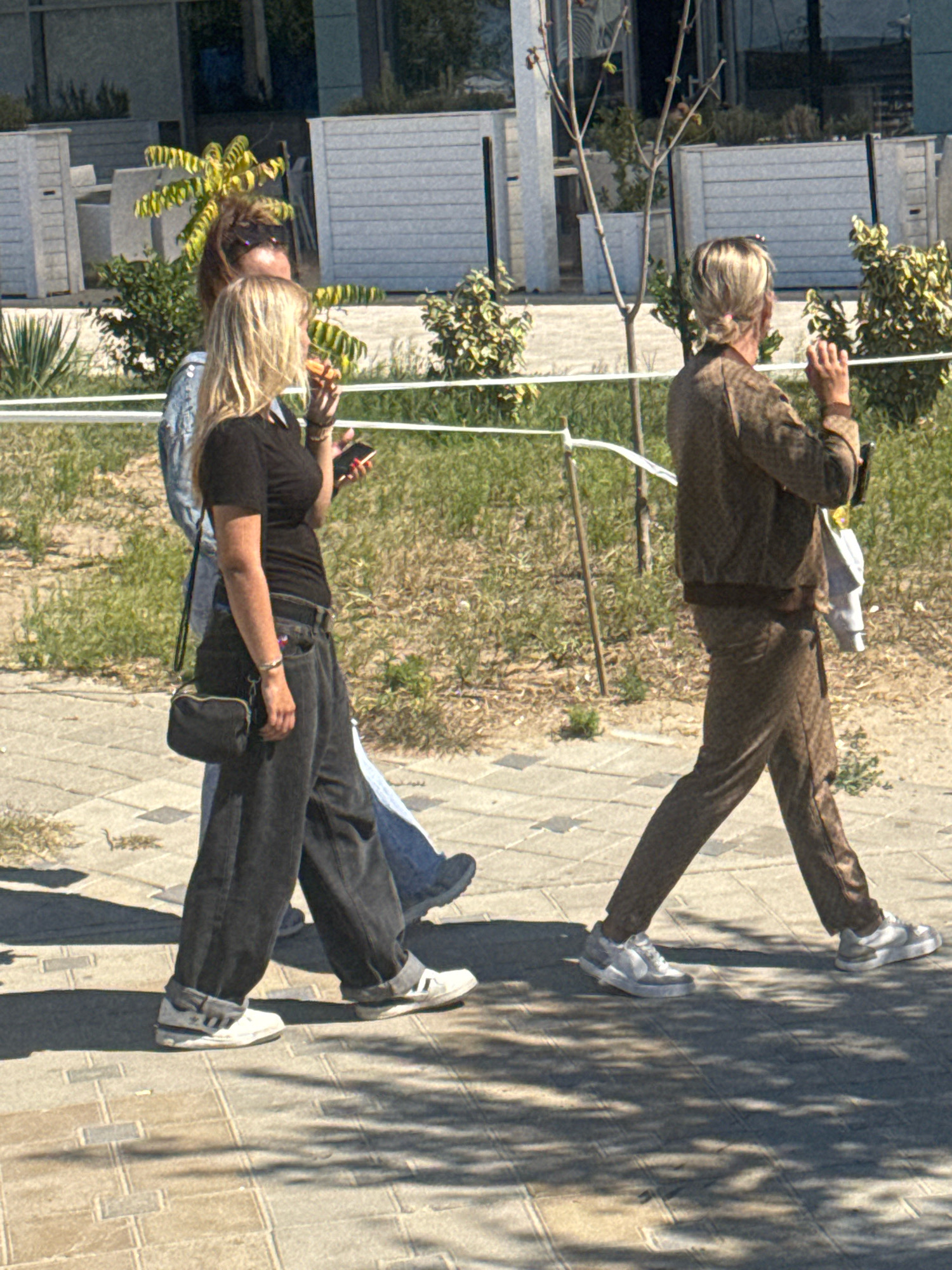 In Anapa, Russia, on a sunny day, three individuals walk along a paved pathway. The foreground features two women and a third person partially visible behind them. The woman closest to the viewer on the left, with blonde hair, wears a black t-shirt and dark, baggy jeans. She carries a small black crossbody bag and holds a mobile phone in her right hand. Next to her, a second woman with blonde hair, seen from the side, wears a denim jacket and holds a piece of food. Behind them, the third individual is mostly obscured but appears to be wearing blue jeans. Further to the right, a woman with blonde hair pulled back walks away from the viewer. She is dressed in a brown patterned tracksuit, consisting of a zipped jacket and matching pants. She carries a light-colored garment over her left arm and holds her right hand up near her face. The environment consists of a paved walkway with shadows of trees cast upon it, bordered by a grassy area with some bushes and trees. In the background, there are modern white structures with parallel lines, possibly part of a building or fencing. White ropes supported by poles run horizontally across the scene, delineating the area.