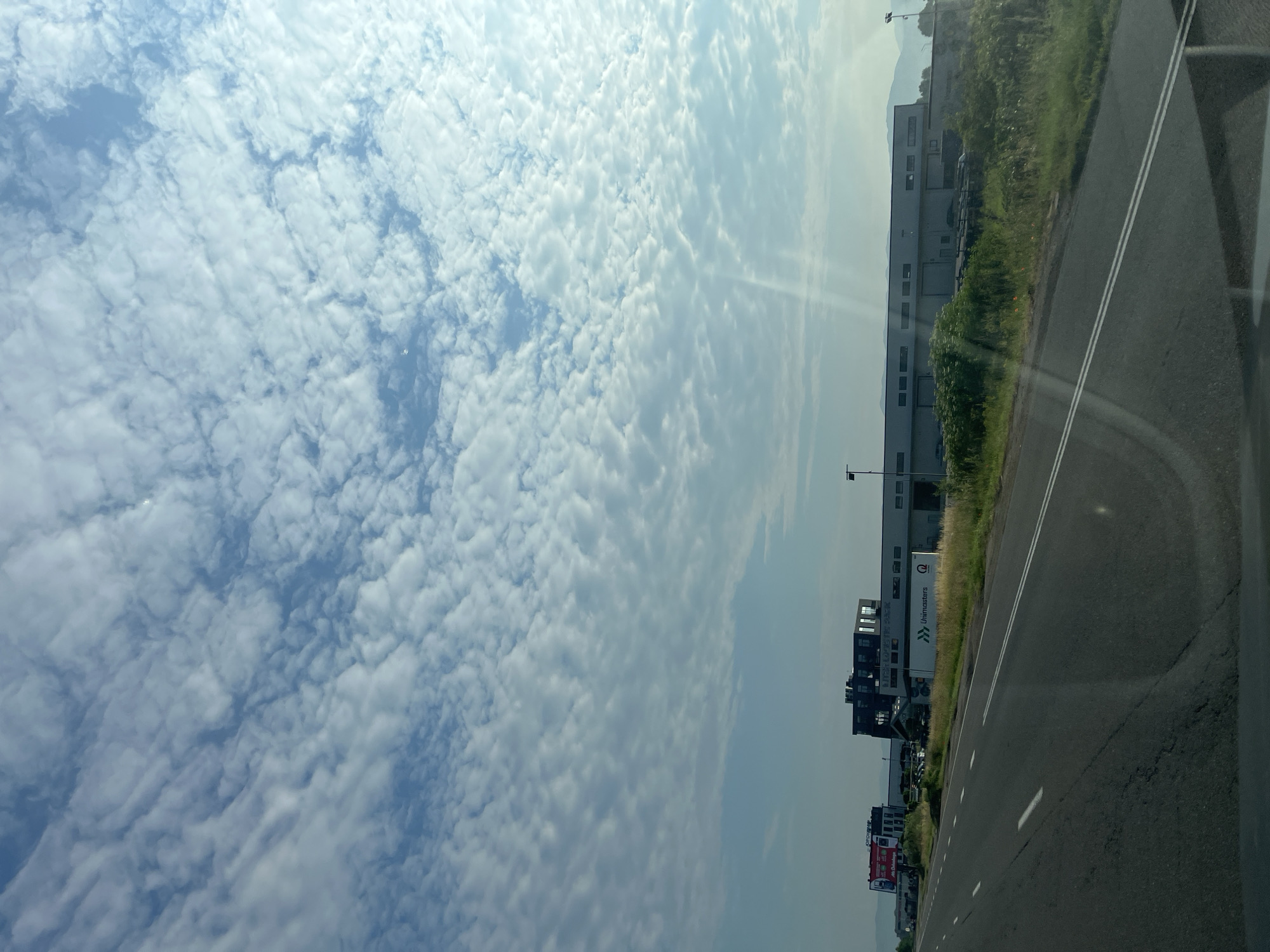 The image depicts a wide, multi-lane road in an outdoor setting under a partly cloudy sky. On the right side of the road, there is a long, industrial building with a white trailer truck parked in front of it, featuring the "Unimasters" logo. Further down the road, on the left, a red and white billboard is visible, along with other buildings. The road itself is paved and marked with white lines. The surrounding environment includes green grass and some trees. The lighting suggests it is daytime.