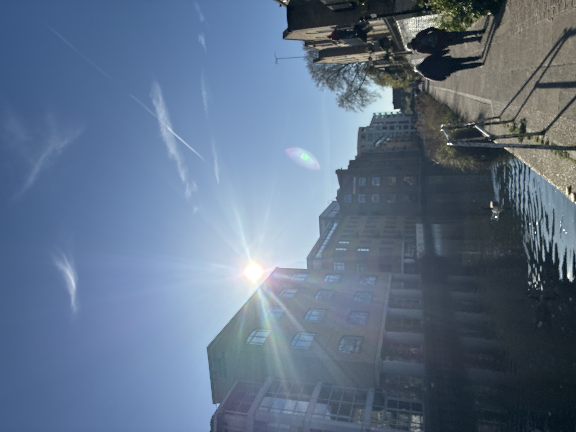 The image depicts a sunny day along a canal in what appears to be London, United Kingdom. The sun shines brightly behind a modern building with numerous windows, creating a strong lens flare. Streaks of clouds are visible in the clear blue sky. Adjacent to the canal is a paved walkway where two figures are walking. A metal railing lines part of the walkway. Other buildings are visible in the background, and there are some trees. The canal reflects the buildings and sky, with some ripples disturbing the reflection.