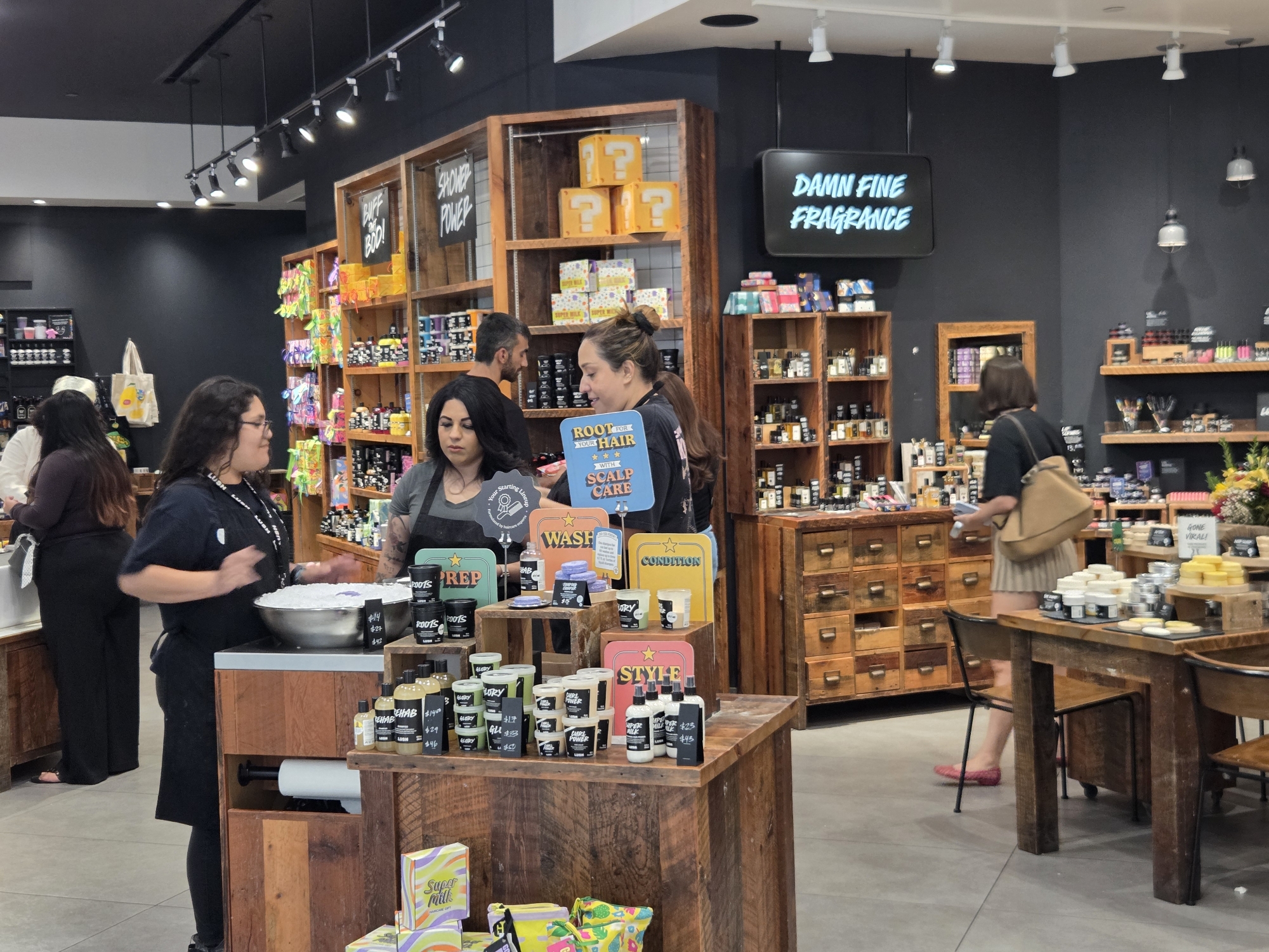 The image shows the interior of a Lush cosmetics store in Glendale, United States of America. The store is brightly lit with overhead track lighting. In the foreground, a woman wearing a black apron and black leggings is standing behind a wooden counter with a large metal bowl filled with white material, likely product ingredients. She is holding a white scoop. To her right, on the counter, are various products in clear bottles and black containers with price tags. Behind her, shelves are filled with more products, including some in colorful packaging.
Further back in the store, other customers and employees are visible. A woman with long dark hair is looking at products on a shelf. A man is standing near some shelves displaying yellow boxes with question marks on them, possibly a themed display. Another woman is browsing near a wooden cabinet with many small drawers. The walls of the store are painted dark gray, and shelves are made of wood, showcasing a variety of Lush products. A digital screen on the wall displays the text "DAMN FINE FRAGRANCE" in glowing blue letters. A wooden table with chairs is visible to the right, with more products displayed on it. The overall impression is of a busy retail environment stocked with cosmetics.