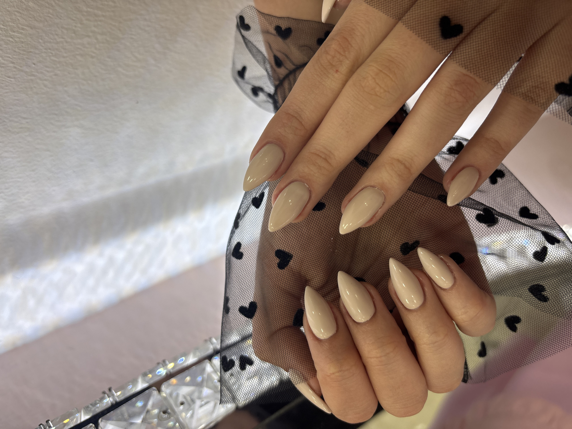 The close-up image features a person's hands, likely female, showcasing a manicure and a unique accessory. Both hands display long, almond-shaped nails coated in a glossy, light beige or nude nail polish. A sheer black fabric, patterned with numerous small black hearts, partially covers the hands and wrists, appearing as a stylish glove or sleeve with a fine mesh texture. In the lower left corner, a highly reflective, faceted object, possibly a crystal bracelet or a decorative element from a bag, catches the light, creating rainbow-like glints. The hands are posed elegantly against a soft, out-of-focus background with varying shades of light beige and pink. The lighting is bright and consistent, indicating an indoor setting. The overall aesthetic suggests a focus on beauty, fashion, and feminine style. There is no visible text in the image. This scene is set in Sofia, Bulgaria.
