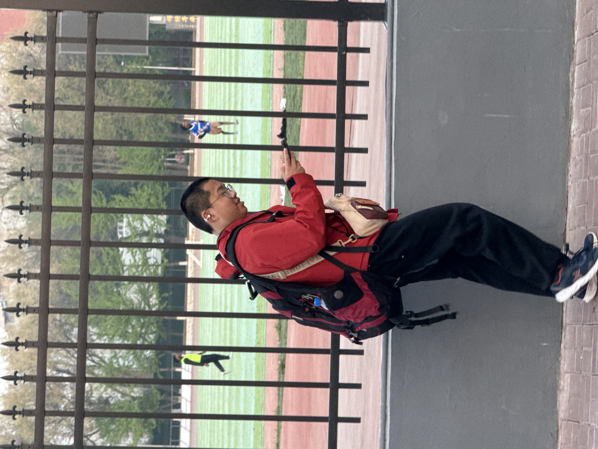 A person wearing glasses, a red jacket, black pants, and athletic shoes stands on a tiled walkway, holding a dark object (likely a phone) at eye level. They also wear a backpack and a smaller bag over their shoulder. Behind them is a dark metal fence with decorative spear-shaped finials. Through the fence, a sports field is visible, with a green grass area, a reddish-brown running track, and two figures on the field: one in a blue uniform and one in a yellow vest. The city of Zhongtun is not explicitly visible in the image.