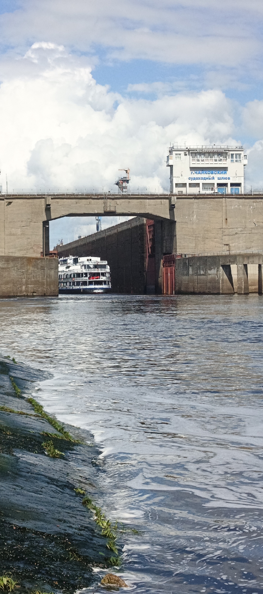 A medium, eye-level shot shows a passenger boat going through the concrete canal of the navigational locks, located in the city of Chaykovsky, Russia. The river water is the foreground, and a low, dark bank is on the left side of the image. The canal is made of gray concrete. The main part of the canal has a section cut out in the middle for boats to pass through. The canal has a building connected to it, and it is a white building with several windows. The words "G. Chaykovsky" are written on the top of the building in Russian. The top right side of the frame is blue sky and white, fluffy clouds.