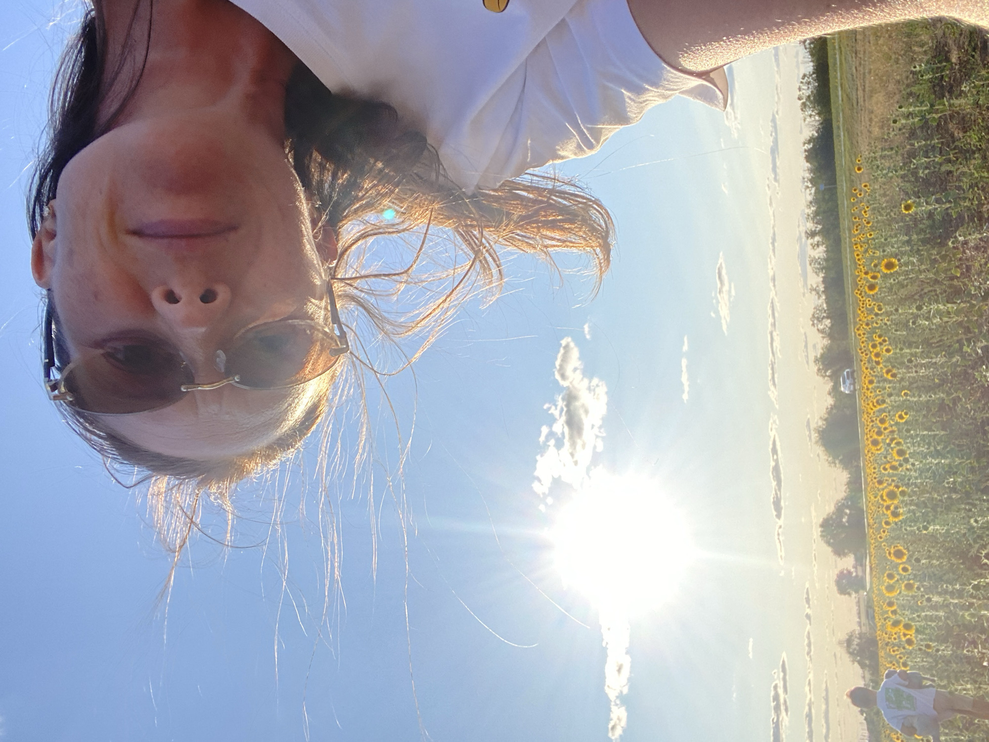 A low-angle selfie captures a person in sunglasses and a white t-shirt, with their face partially obscured by the bright sun and windblown hair, standing in a vast sunflower field. The sun is a strong, bright orb in the blue sky, casting a lens flare across the image. In the distance, a field of sunflowers stretches towards a tree line and a road with a white car. A small child, facing away from the camera, is visible in the left portion of the sunflower field. The scene appears to be set in a rural area.