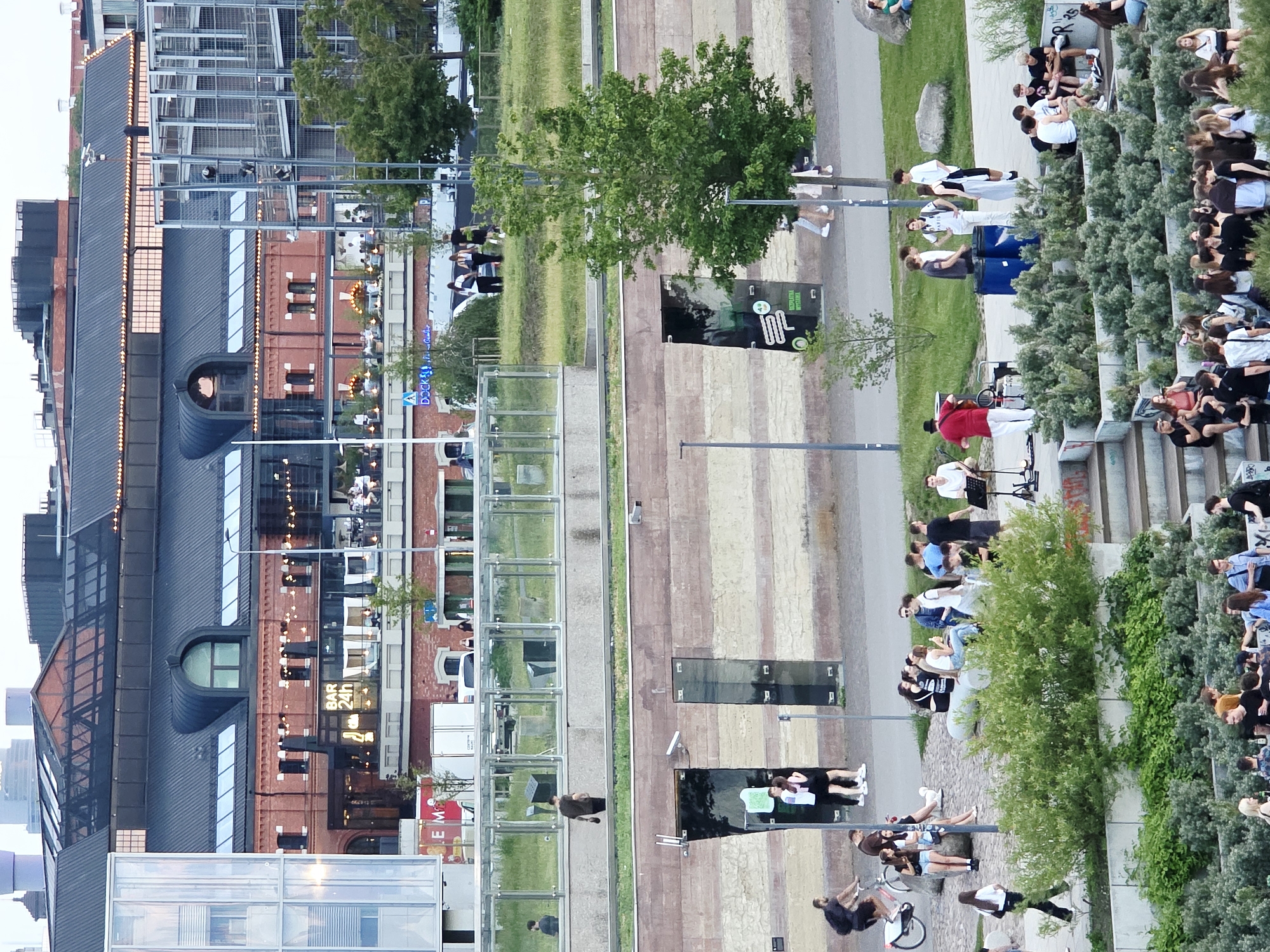 The image shows a scene in Warsaw, Poland, likely near the Vistula River. The perspective is from an elevated viewpoint, looking down upon a public space with several levels. In the background, a distinctive red brick building with a modern black roofline is visible, featuring large windows and an upper-level outdoor seating area with umbrellas and lights. A prominent sign on this building reads "BAR 24h". To the left, a modern glass and steel structure is present.
In the mid-ground, a wide, paved walkway runs along a wall constructed of horizontal bands of light and dark brick or stone. Two doorways with glass fronts are set into this wall. People are walking and standing on this walkway.
The foreground is dominated by a green, terraced park-like area with abundant vegetation and stone seating. A group of people are gathered in this area, some sitting on the stone structures and others standing. One group appears to be watching a musical performance by a man in a red shirt and hat, who is playing an instrument and accompanied by two blue barrels. Other individuals are scattered throughout the scene, engaged in various activities such as cycling, walking, and conversing. The overall atmosphere appears to be a casual, outdoor gathering in an urban setting.