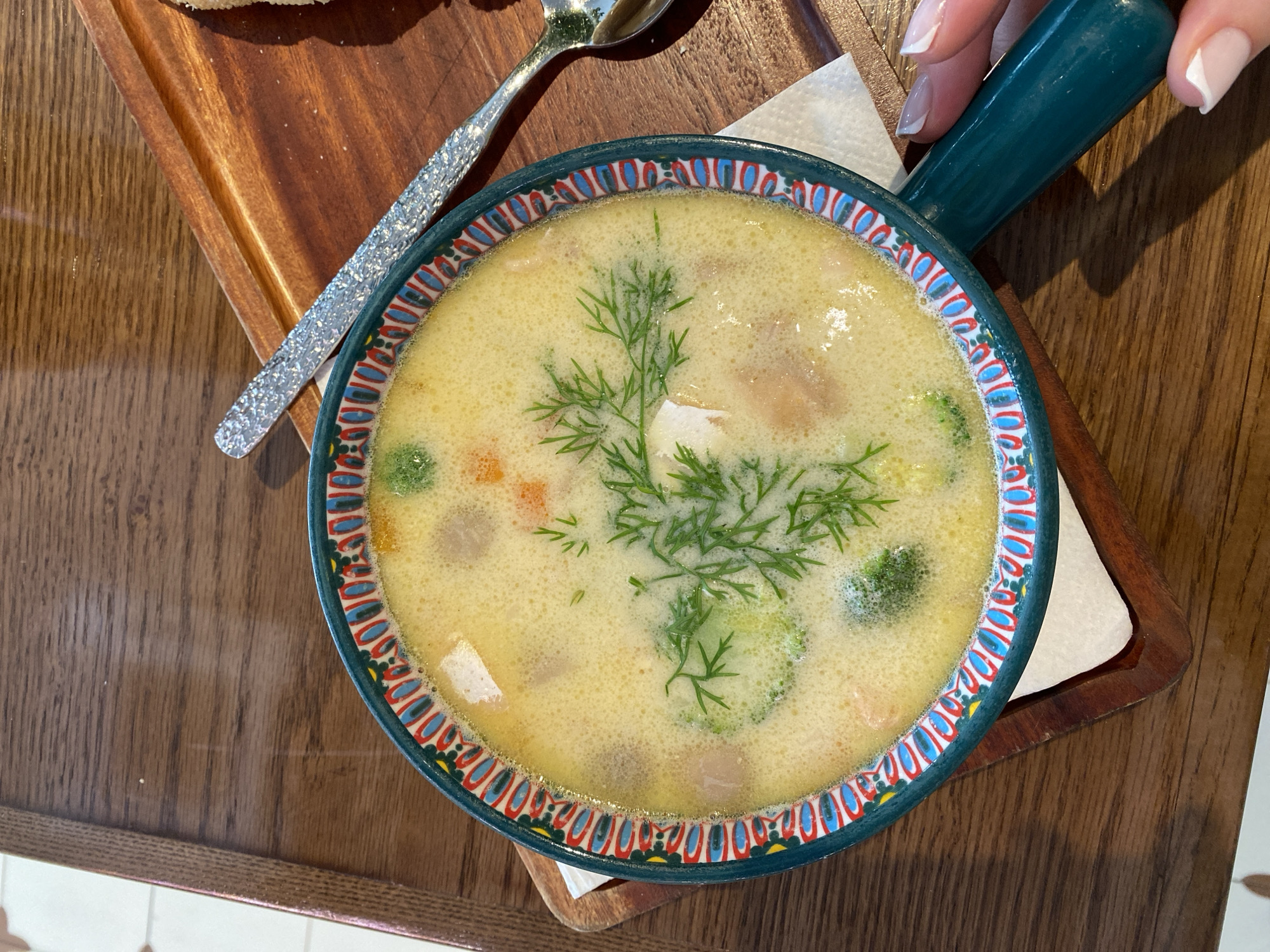 A close-up overhead shot shows a bowl of creamy soup with dill, broccoli, and chunks of fish or chicken. The bowl has a decorative rim with a pattern of blue, red, and yellow. It rests on a wooden serving tray, which is on a wooden table. A spoon with a textured handle is placed on the tray. A person's hand with manicured nails is holding the bowl's handle. The background shows a tiled floor and the edge of a wooden table. The image does not provide any information that can be used to determine its location in Đà Nẵng, Vietnam.