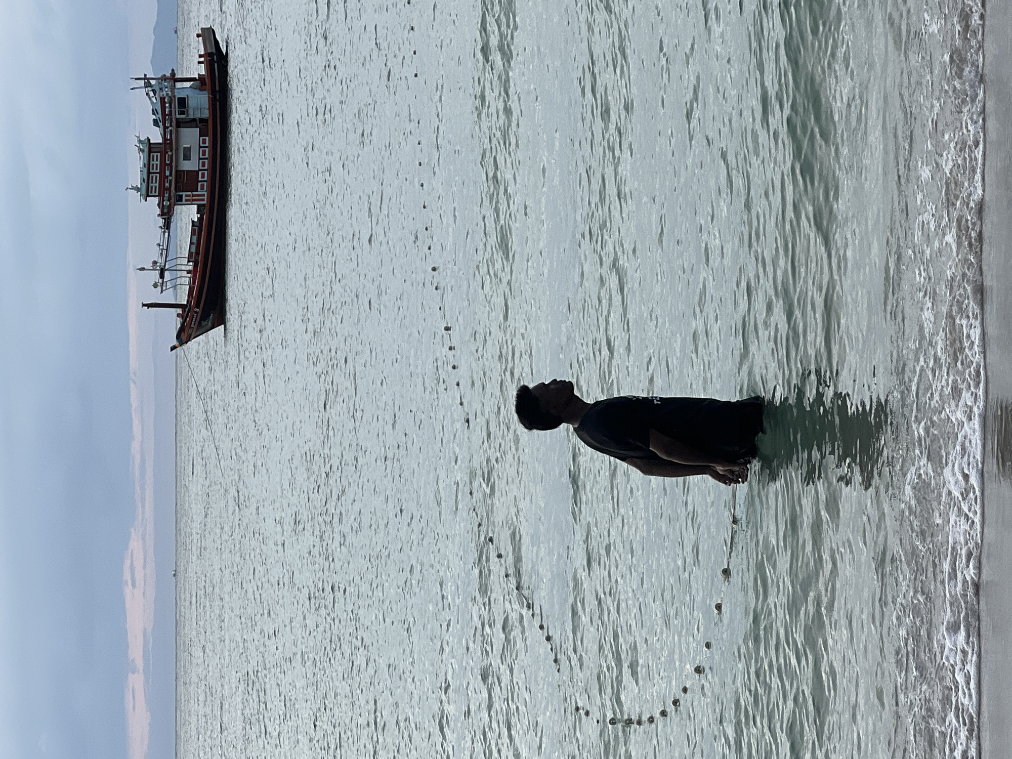 A lone man stands in shallow ocean water, facing to his right. He is wearing a black t-shirt and his arms are behind his back. In the background, a red and white fishing boat is anchored in the water. A string of buoys stretches across the water in front of the man and extends to the right. The sky is overcast, and the water is calm. The scene appears to be on a beach with gentle waves lapping at the shore.