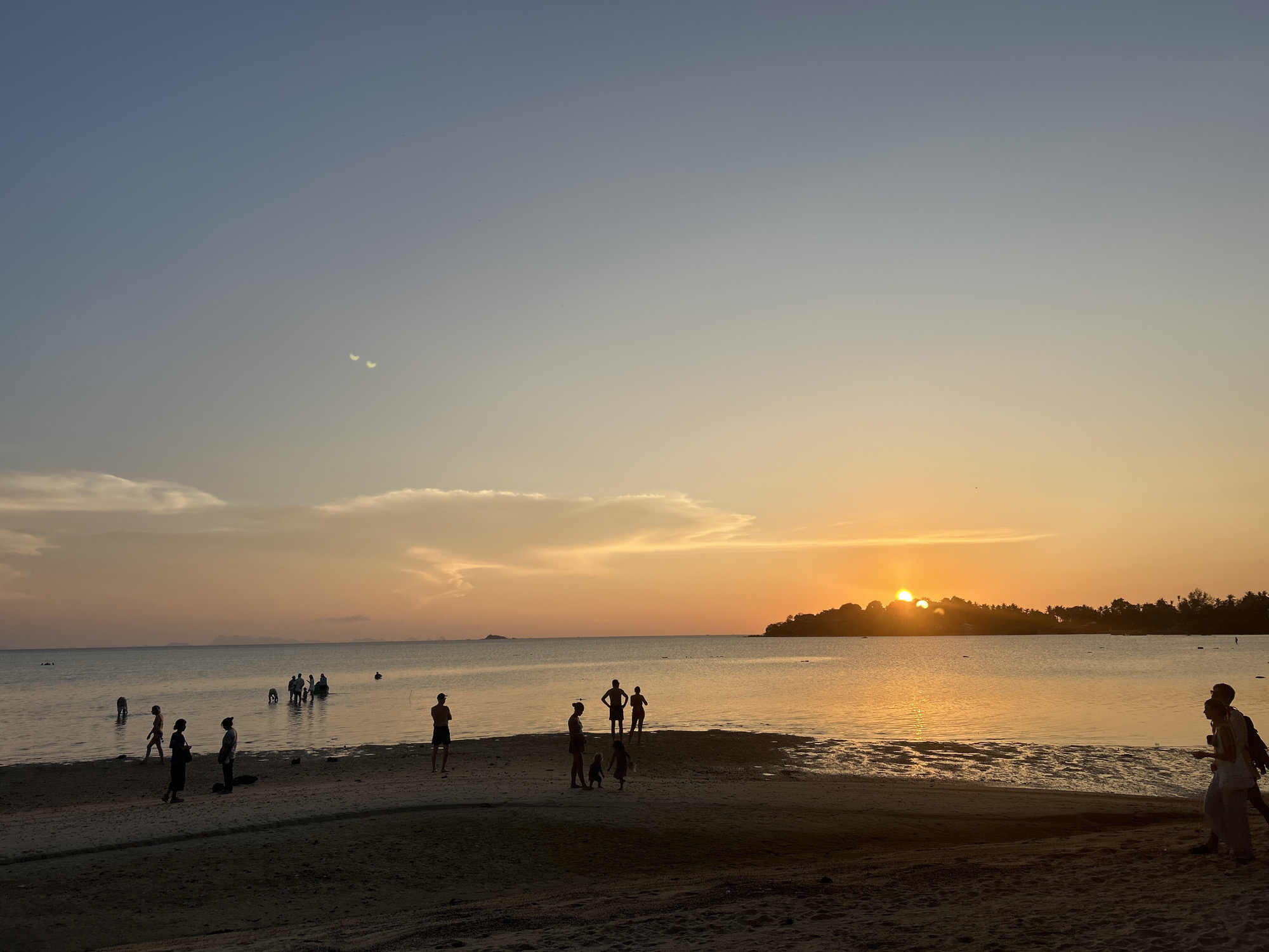 A photograph captures a sunset scene at Baan Sri Thanu, Thailand. The sun is positioned near the horizon behind a land formation, casting a golden hue on the water's surface. Several groups of people are visible along the beach, some standing on the sand while others are wading in the water. These people are silhouetted against the bright sky, indicating they are standing between the viewer and the light source. The sky transitions from blue at the top to a gradient of orange and yellow closer to the horizon, marked by some cloud cover. The foreground consists of a sandy beach, with distinct textures and patterns.