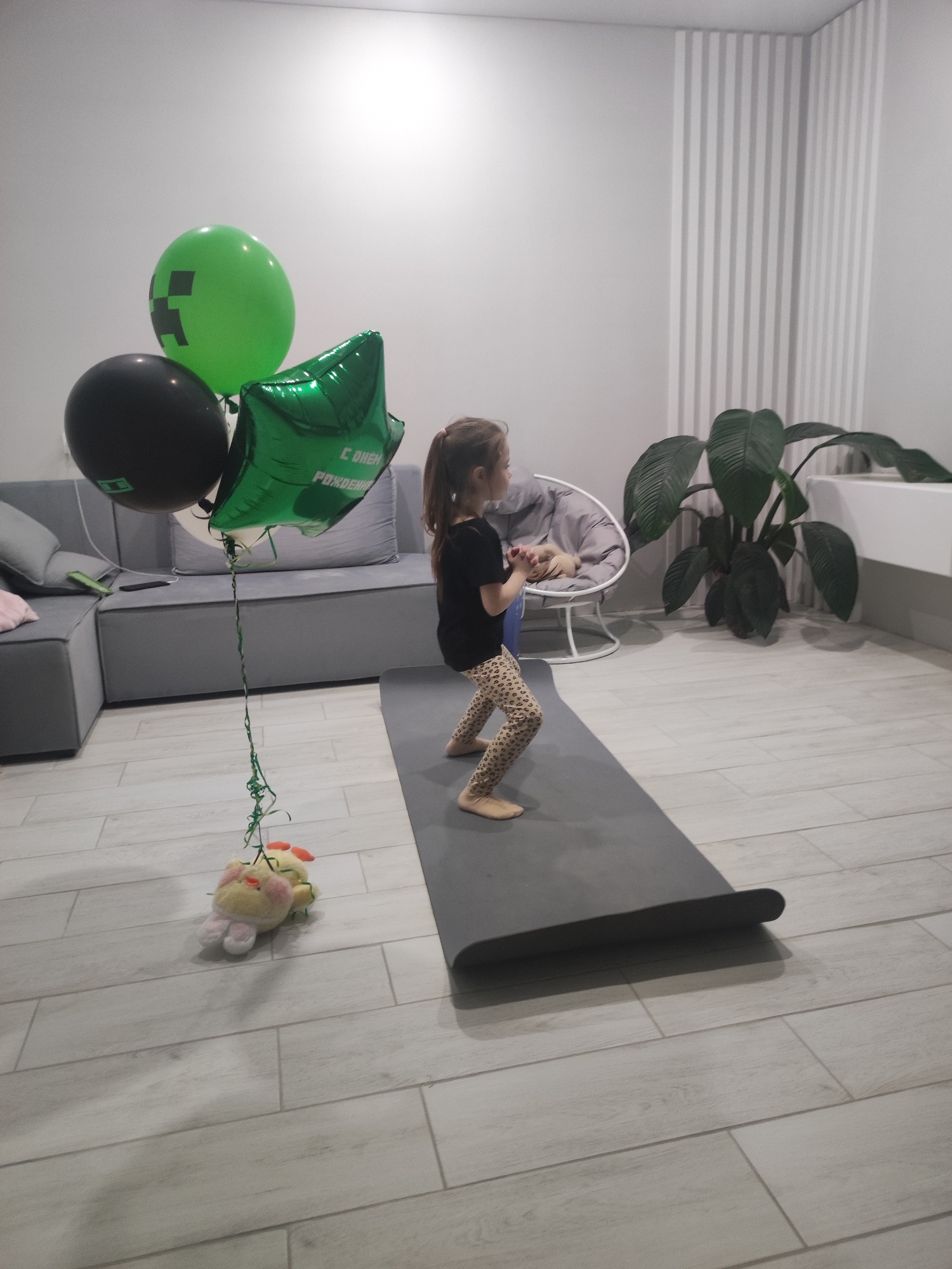 The image shows a young girl in a brightly lit room. She is wearing a black t-shirt and patterned leggings, standing on a gray yoga mat. A bouquet of balloons, including a green Minecraft-themed balloon, a black balloon, and a green star-shaped balloon with Russian text, is attached to a stuffed animal on the floor. The balloons are positioned near a gray sectional sofa. The room has a modern interior with light gray walls and tiled floors. There is a decorative accent wall with vertical stripes, a large potted plant, and a round chair in the background. The scene suggests a celebration or birthday event. There is no information to suggest that this image was taken in Votkinsk, Russia.