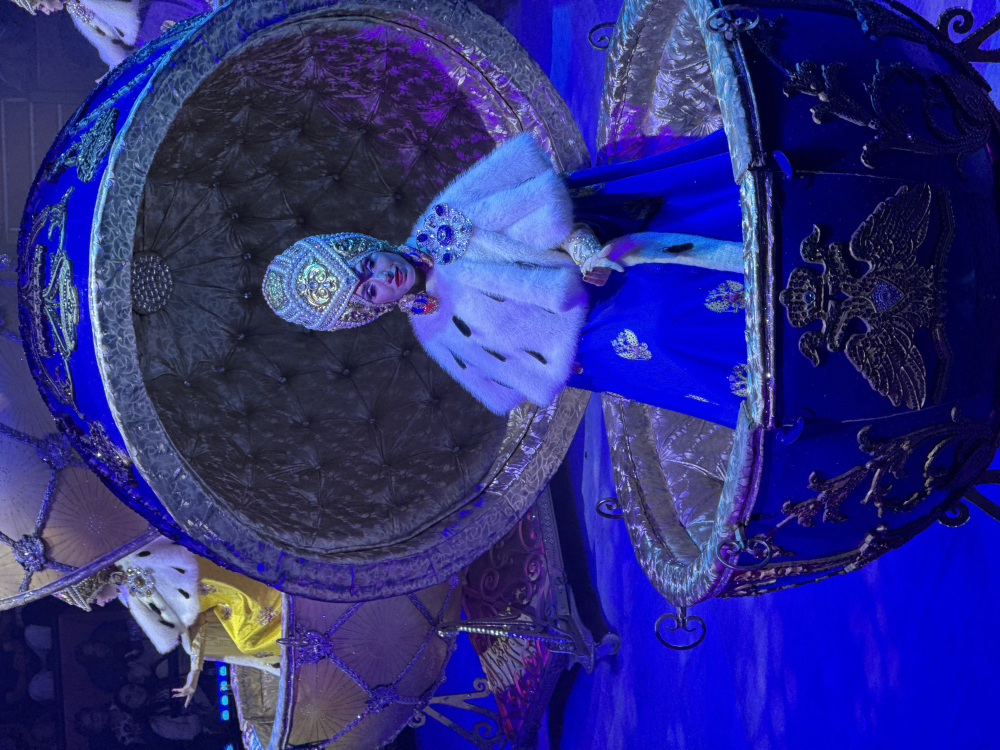 A woman in a bejeweled kokoshnik headdress and a white fur-trimmed cloak over a blue dress sits in a gilded, egg-shaped carriage on a blue stage. She is in the foreground, facing the camera with her left hand extended slightly. Behind her, another similar carriage with a woman in a yellow dress is partially visible. The carriages are ornate, with elaborate golden detailing. The lighting is predominantly blue and purple, suggesting a theatrical performance or show. The background shows an audience in dim lighting. The setting is the city of Sochi, Russia.