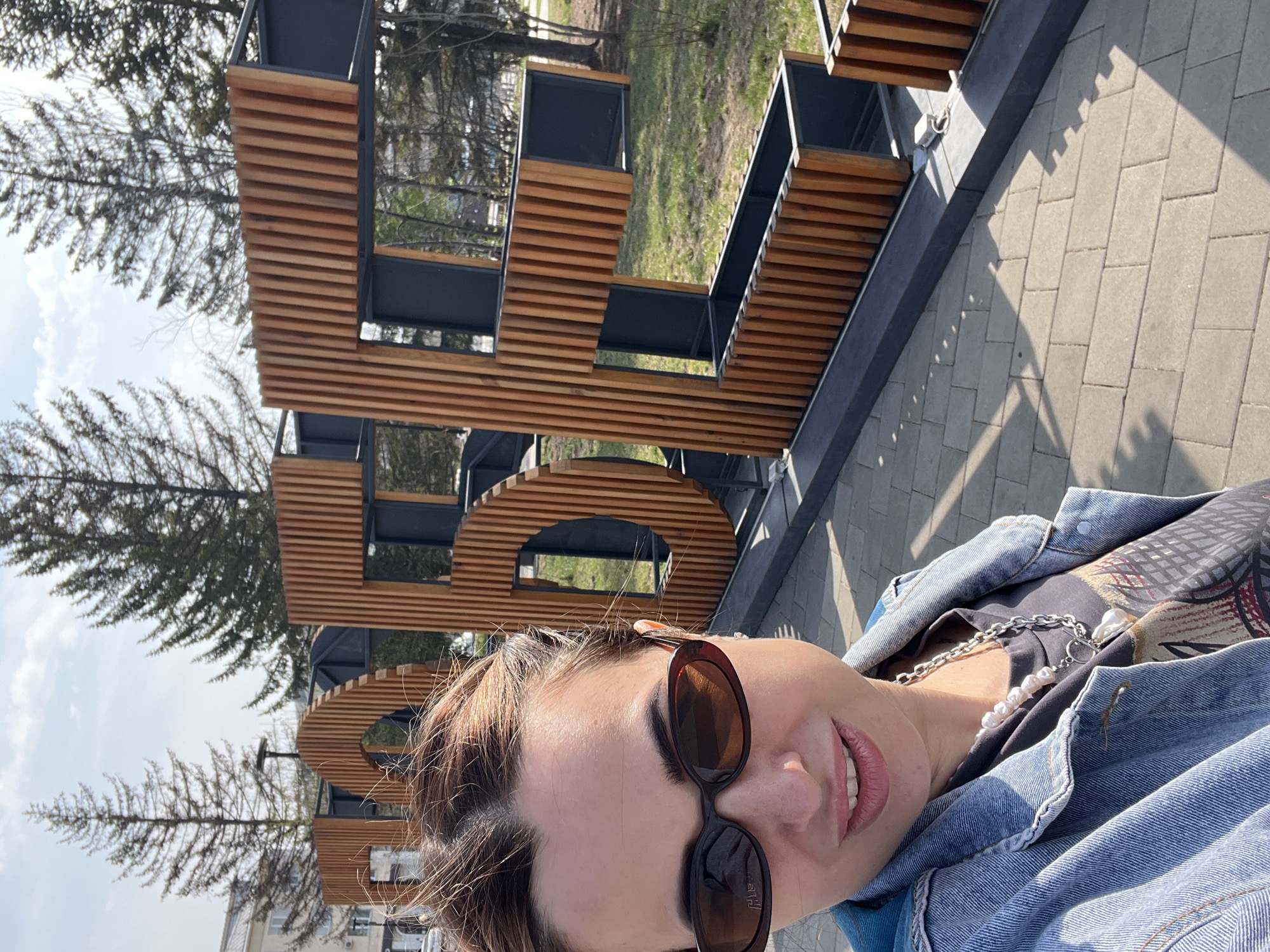 A medium-shot selfie of a woman wearing sunglasses and a denim jacket, taken outdoors on a sunny day in a city of Unknown, Unknown. She is standing in front of a large, wooden, open-lettered structure, possibly spelling out a word or name of the location. The structure is positioned on a paved area. The background includes trees and buildings.