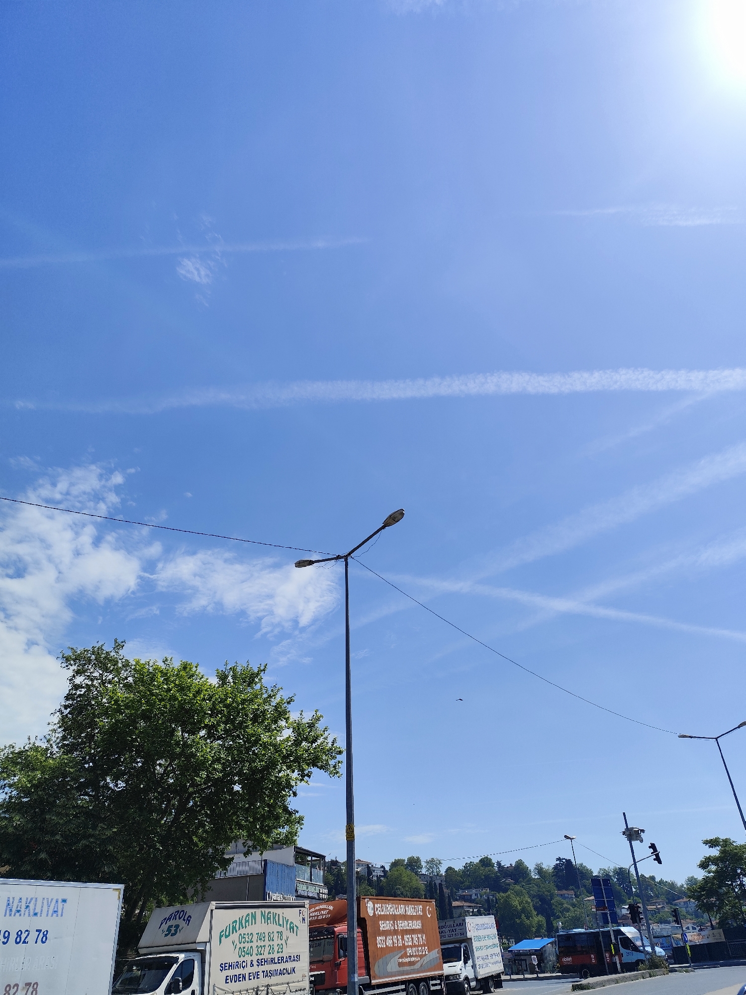 This media file captures a bright daytime street scene in Sarıyer, Turkey, featuring a vivid blue sky adorned with several straight contrails and scattered wispy clouds. The sun's glare is prominent in the upper right corner.
In the foreground, large green trees with dense foliage partially obscure the background on the left. Several white and orange moving trucks are visible, parked or driving along a road. Prominently displayed text on these trucks, written in Turkish, indicates they are "NAKLIYAT" (transportation/moving) companies, specializing in "ŞEHİRİÇİ & ŞEHİRLERARASI" (intercity & intracity) and "EVDEN EVE TAŞIMACILIK" (house to house moving). Specific company names like "FURKAN NAKLİYAT" and "ÇELİKOĞULLARI NAKLİYAT" are readable, along with various phone numbers (e.g., 0532 749 82 78, 0540 327 28 29, 0532 466 19 26, 0535 745 76, 212 323 53 52). One truck also shows "PAROLA 53".
A tall street light pole with an attached lamp stands in the mid-ground, with utility lines crossing the frame. In the background, a hillside covered with green trees and numerous buildings is visible under the clear sky. Another small vehicle, possibly a bus, with "obilet" written on its side, is also seen further down the road. The overall impression is a busy, sunny day in an urban or suburban area.