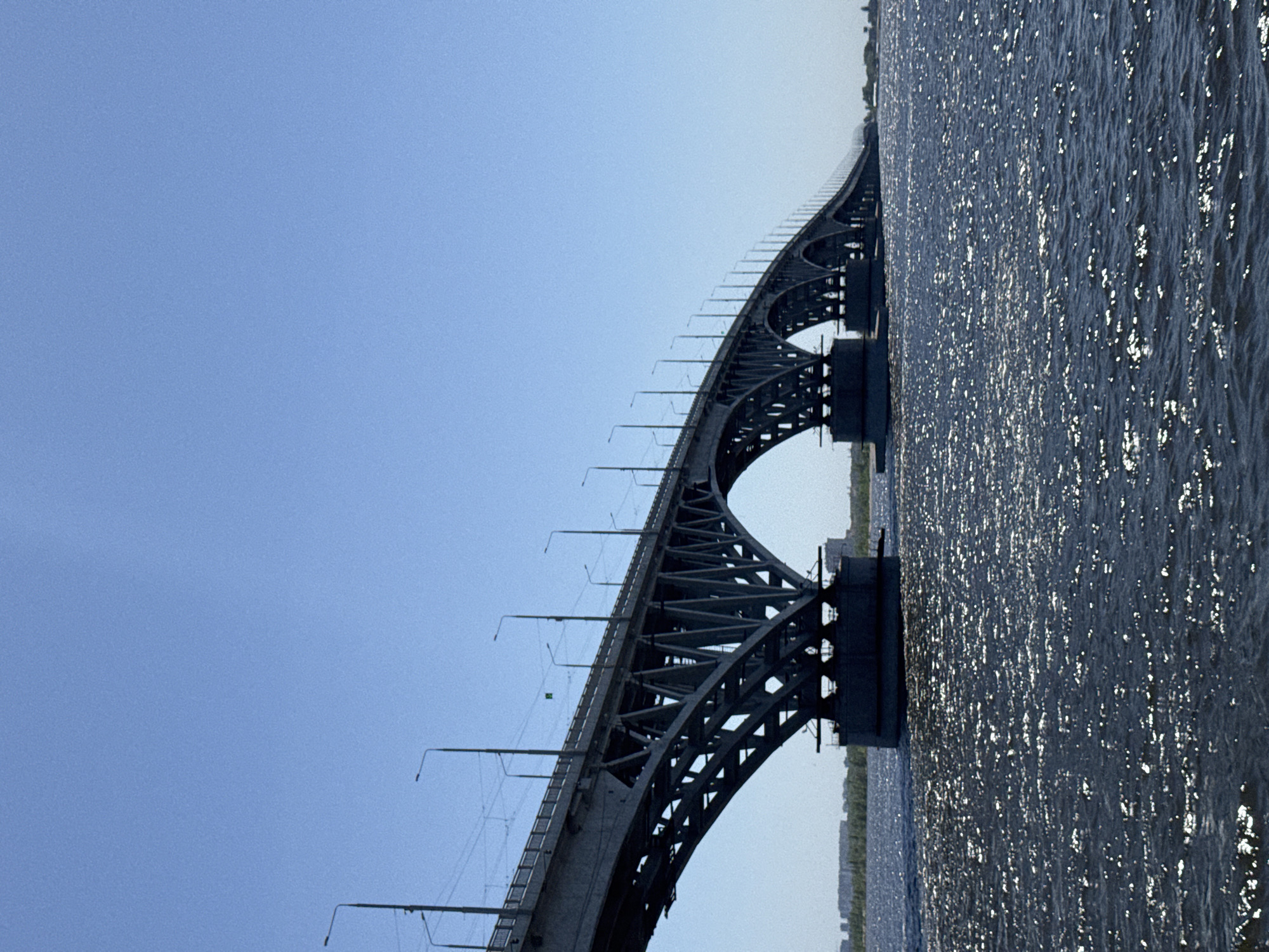 The image depicts the Saratov Bridge spanning the Volga River in Saratov, Russia. The bridge is a large, arched steel structure extending horizontally across the frame. The left side of the image shows a clear, blue sky. The right side captures the Volga River with light reflecting on its surface, creating a shimmering effect. Vegetation lines the banks in the distance. The bridge itself features overhead wires, suggesting it may carry rail traffic in addition to road vehicles.