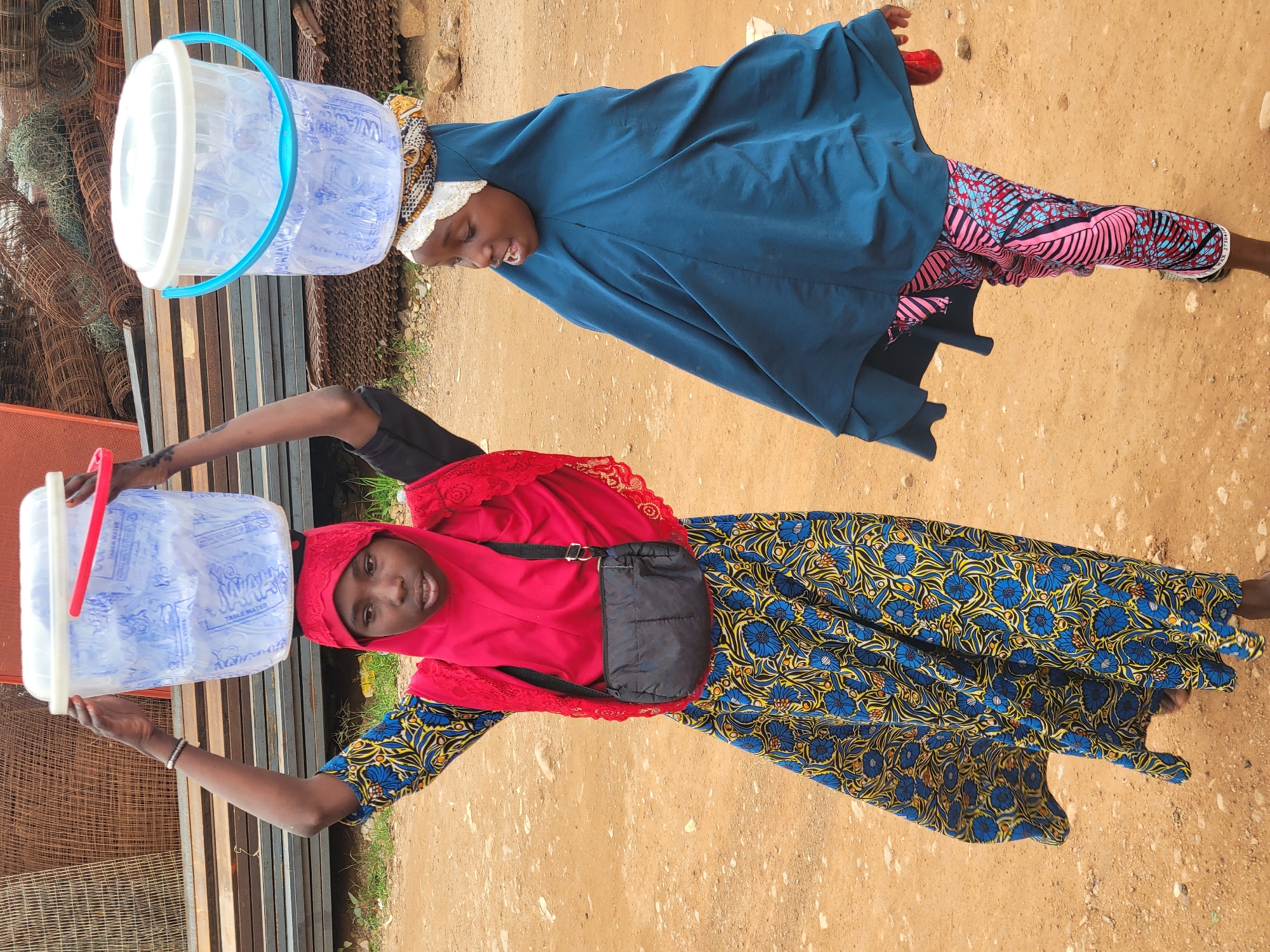 Two young girls in traditional clothing are walking side-by-side, each carrying a large bucket of water balanced on their heads. The girl in the foreground wears a red headscarf and a vibrant blue and yellow floral patterned dress. She has a small black bag slung across her chest. The girl behind her wears a dark blue head covering and a pink and blue patterned fabric on her lower body. Both girls appear to be looking forward, suggesting they are on a journey or heading to a destination.
The setting is outdoors, with a dusty, sandy ground. In the background, there are stacks of metal bars and what appears to be fencing material, indicating a possibly industrial or construction-related area. Some greenery is visible on the right side. The lighting suggests it is daytime, likely mid-morning or afternoon, with clear weather. The overall atmosphere seems focused and determined, with the girls concentrating on their task of carrying the water. The buckets are transparent, filled with water, and have blue handles. Visible text on the buckets reads "WATER TABLE WATER". The image captures a moment of daily life and labor.