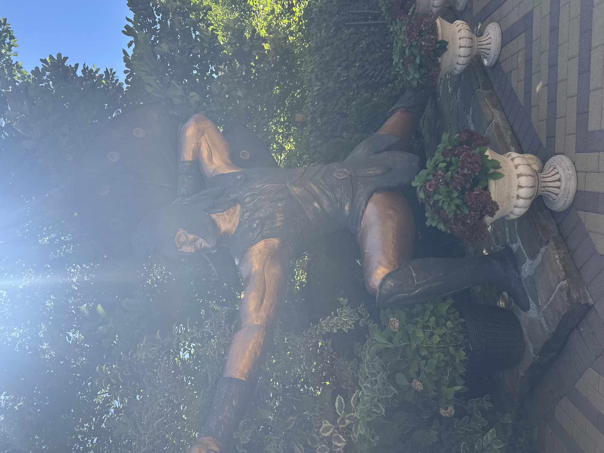 A full-body bronze statue of a muscular warrior in a helmet and armor is positioned outdoors, set against a backdrop of lush green foliage and a bright blue sky. The warrior is depicted in a dynamic pose, with one arm raised and holding a large, circular shield over its head, while the other arm is extended forward, as if aiming a weapon. The statue's metallic surface has a warm, coppery hue, and it is partially obscured by the dappled sunlight filtering through the leaves.
In the foreground, the statue stands on a raised platform constructed from interlocking brick pavers and stone slabs. Flanking the platform are two ornate, white urns filled with dark red flowering plants. To the left of the statue, a dark woven basket holds a green leafy plant. The overall setting suggests an outdoor garden or decorative area, possibly a public space within Krasnaya Polyana, Russia, given the context. The sunlight creates lens flare and highlights, adding a bright and somewhat ethereal quality to the scene.