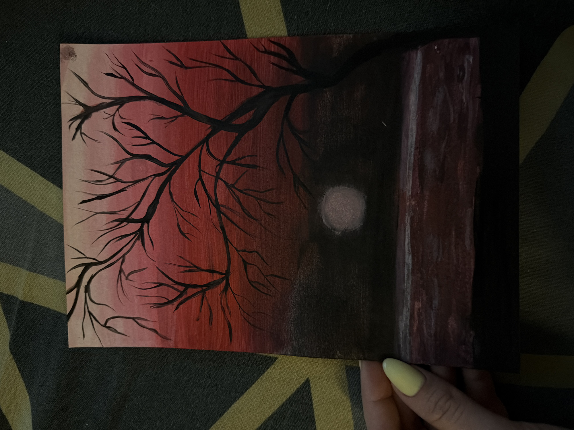 A painting is held by a hand with a pale yellow nail. The painting depicts a tree with bare branches against a red and black sky. A circular, white object resembling a moon is visible in the darker portion of the sky. A maroon rectangular shape sits on the right edge of the painting. The painting sits on a fabric surface. The location in Murino, Russia is undetermined.