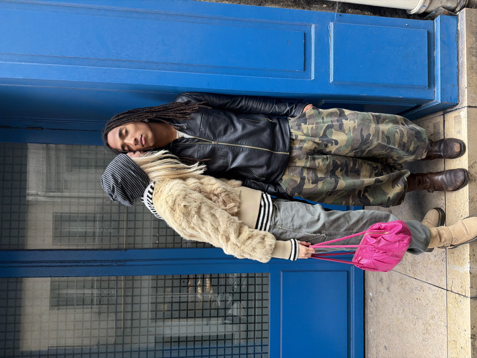 Two individuals are positioned on a light-colored stone sidewalk against a vibrant blue door and adjacent building structure in an urban setting, consistent with Paris, France. No specific address or landmark is identifiable.
The individual on the left has blonde hair, wears a black and white striped beanie, a tan faux fur bomber jacket with striped knit cuffs and hem, grey baggy pants, and light brown suede boots. A fuchsia pink handbag is held in their left hand. This individual is leaning inward, kissing the right cheek of the second individual.
The individual on the right has dark skin and long dreadlocks. They are wearing a black leather jacket over a light shirt, baggy camouflage print trousers, and dark brown leather boots. This individual's eyes are closed, and they are leaning against the blue door frame with their right hand in a pocket.
The immediate background consists of the vibrant blue door and its frame. To the left, a window pane with a reflective grid pattern partially obscures a view of an urban building. A white downspout or pipe is visible on the far right, adjacent to the blue door frame. The ground is a uniform light-colored stone pavement.