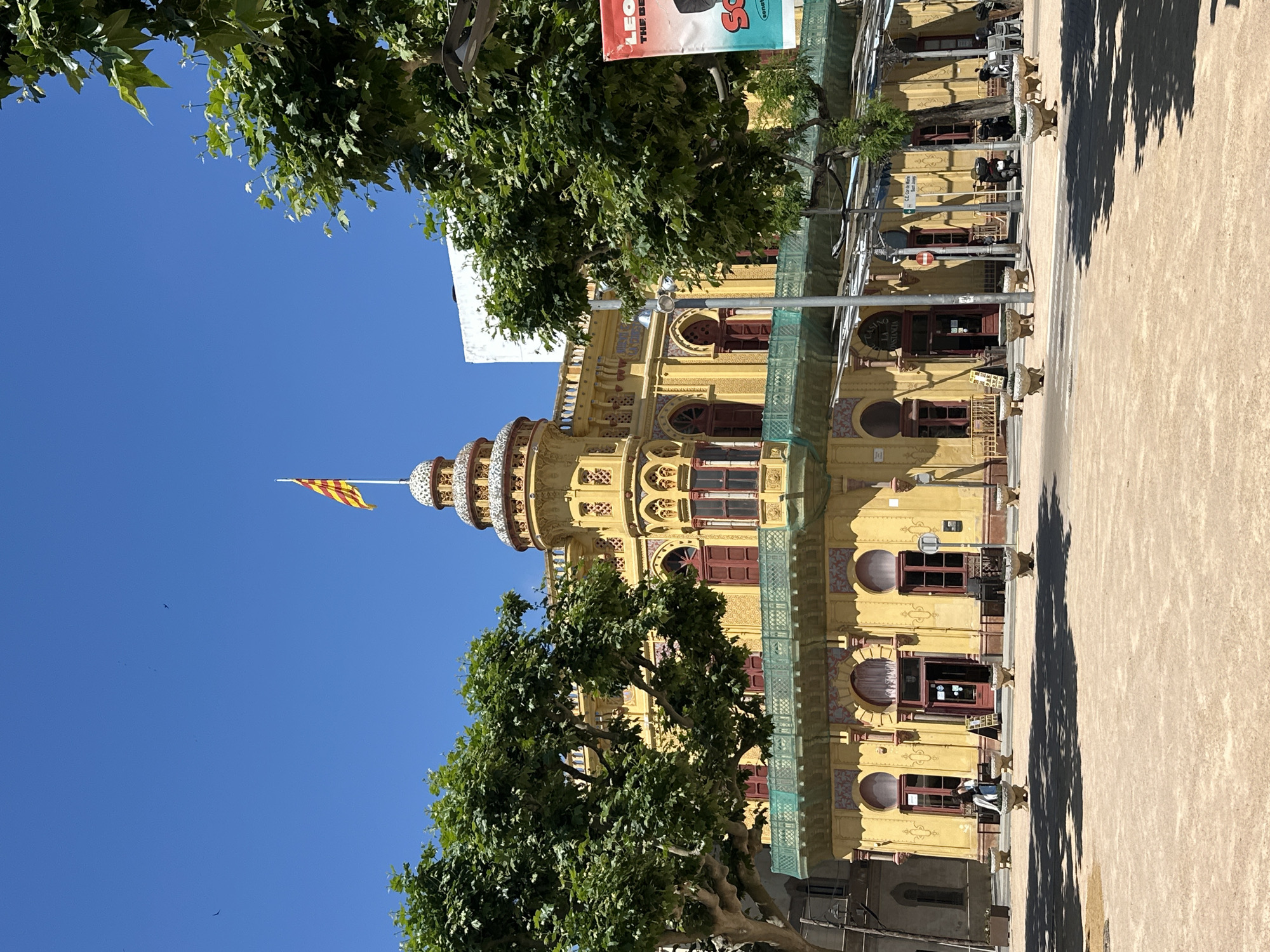 The image features the Casino dels Nois building in Sant Feliu de Guíxols, Spain. The building is yellow with red window frames and intricate decorative details. A flag is visible atop the building's tower against a clear blue sky. Trees are situated in the foreground, partially obscuring the building's facade. A portion of a banner or sign hangs above the building. The ground is light-colored. Pedestrians and parked motorcycles are visible in the background.