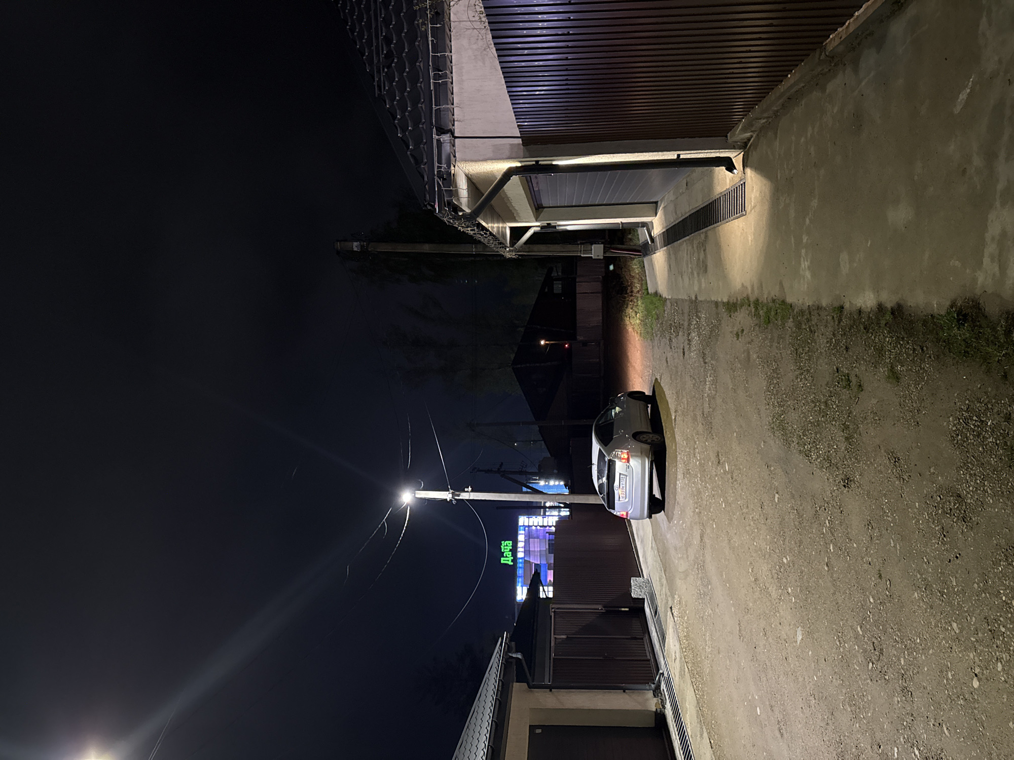 The image depicts a narrow alleyway at night in Молодёжный, Russia. The alley is flanked by buildings on both sides, with a silver car parked on the right side of the frame. A street light illuminates a portion of the alley, revealing a dirt and gravel surface. A neon sign, visible in the distance, spells out "Дача". The sky is dark.