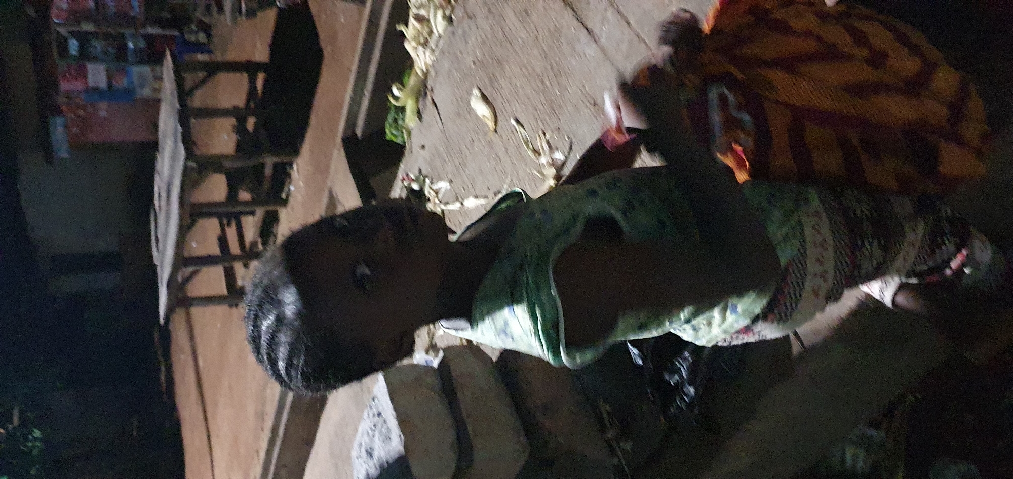 A low-angle, full-body shot captures a young Black child in a dimly lit outdoor setting. The child, facing slightly towards the right, has dark hair styled in braids and is wearing a green and white striped top and patterned pants. They are holding a bundle wrapped in orange and red fabric. To the left of the child are two large, light-colored rectangular objects, possibly stones or bricks. The background shows a rough, brown surface, a wooden table with legs, and what appears to be a building or structure with some colorful signs. Some debris, resembling dried plant material, is scattered on the ground. The lighting is poor, suggesting it is nighttime or a very dark environment. The location is identified as Unknown, Unknown.