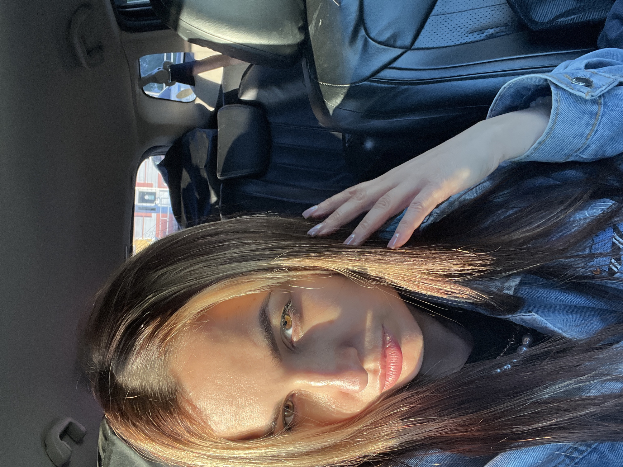 A young woman with light brown hair and blonde highlights is sitting in the passenger seat of a car. She is wearing a denim jacket over a black shirt. Her hand is resting on her head. The car has black leather seats. The location is the city of Unknown, Unknown.