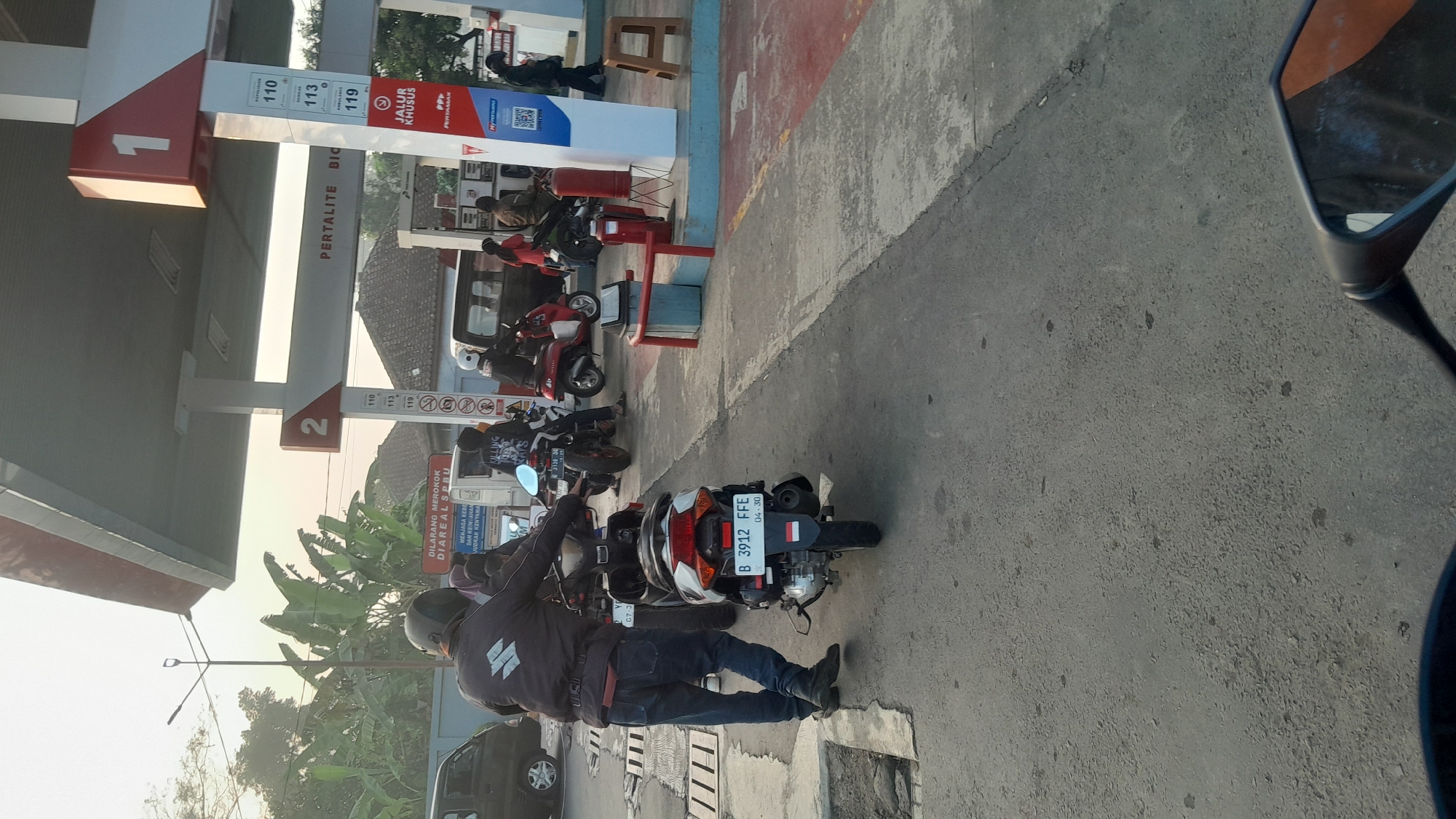 A motorcycle license plate with the text "B 3912 FFE" is visible in the foreground of the image. The license plate also features a small Indonesian flag. The scene appears to be a gas station, with the top of a gas pump visible in the upper right quadrant. Several motorcycles and people are present at the gas station. The environment is outdoors, with a paved surface. The overall lighting suggests it is daytime. The presence of "Pertamina" branding on the gas pump and the style of the license plate are indicative of a location in Indonesia, possibly Subang.