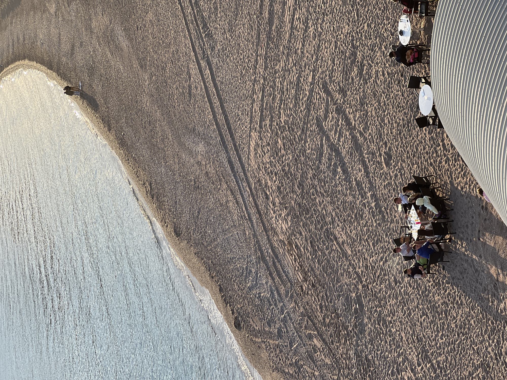 An aerial view looking down on a beach in Budva, Montenegro, shows the Adriatic Sea on the left, with sandy beach comprising the right. A group of people are seated at tables near a corrugated metal structure. Tire tracks are visible in the sand between the waterline and the tables.