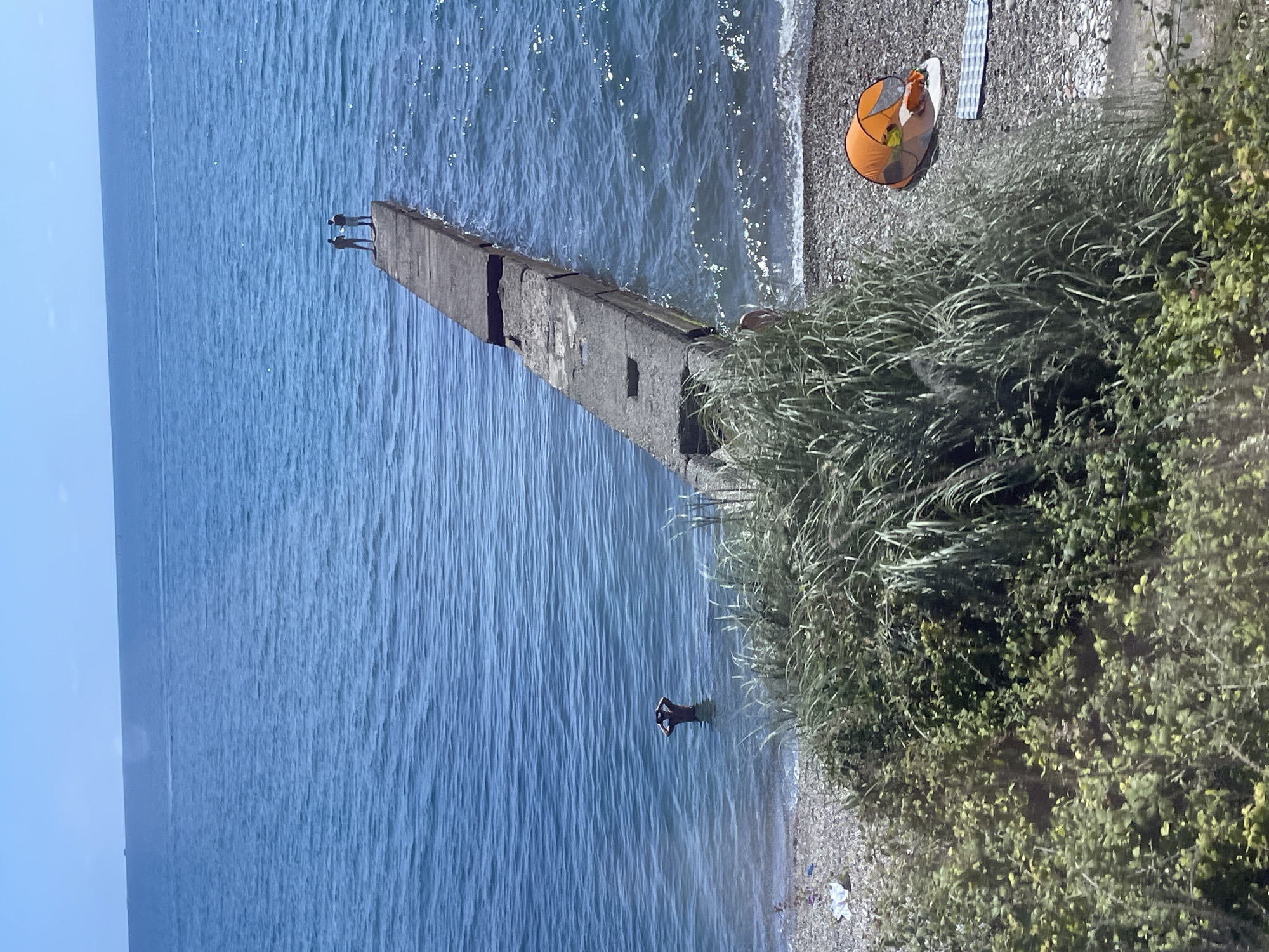 Two individuals are standing on a concrete pier extending into a calm, blue sea. In the shallower water near the shore, a third person is partially submerged, with their hands raised to their head. The beach is comprised of small, grey pebbles and there is lush green vegetation in the foreground obscuring part of the view. An orange tent is set up on the beach, with a patterned towel laid out nearby. The sky is clear and blue, indicating a sunny day. The scene appears to be a coastal area, possibly in Sochi, Russia, given the architectural style of the pier and the general ambiance of a Black Sea resort.