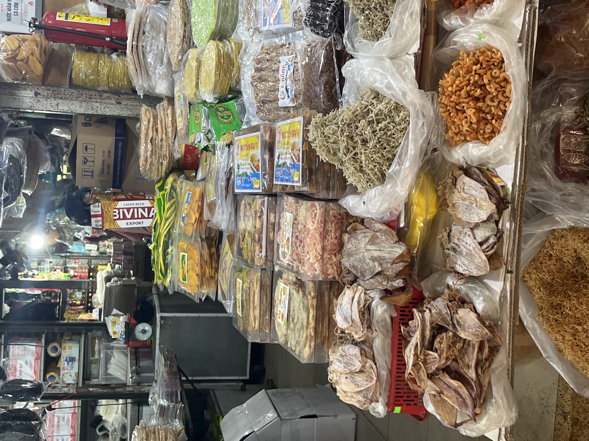 The image shows a stall at a market in Nha Trang, Vietnam. The stall is filled with various dried seafood products, including dried squid, dried anchovies, and dried shrimp. There are also stacks of packaged food items, such as crackers and what appear to be rice paper wrappers. The lighting suggests it is daytime, and the overall atmosphere is that of a busy marketplace. The scene is focused on the merchandise, with no prominent human subjects or actions captured in detail, suggesting a candid or observational approach. The environment appears to be an indoor market or a covered section of an outdoor market.