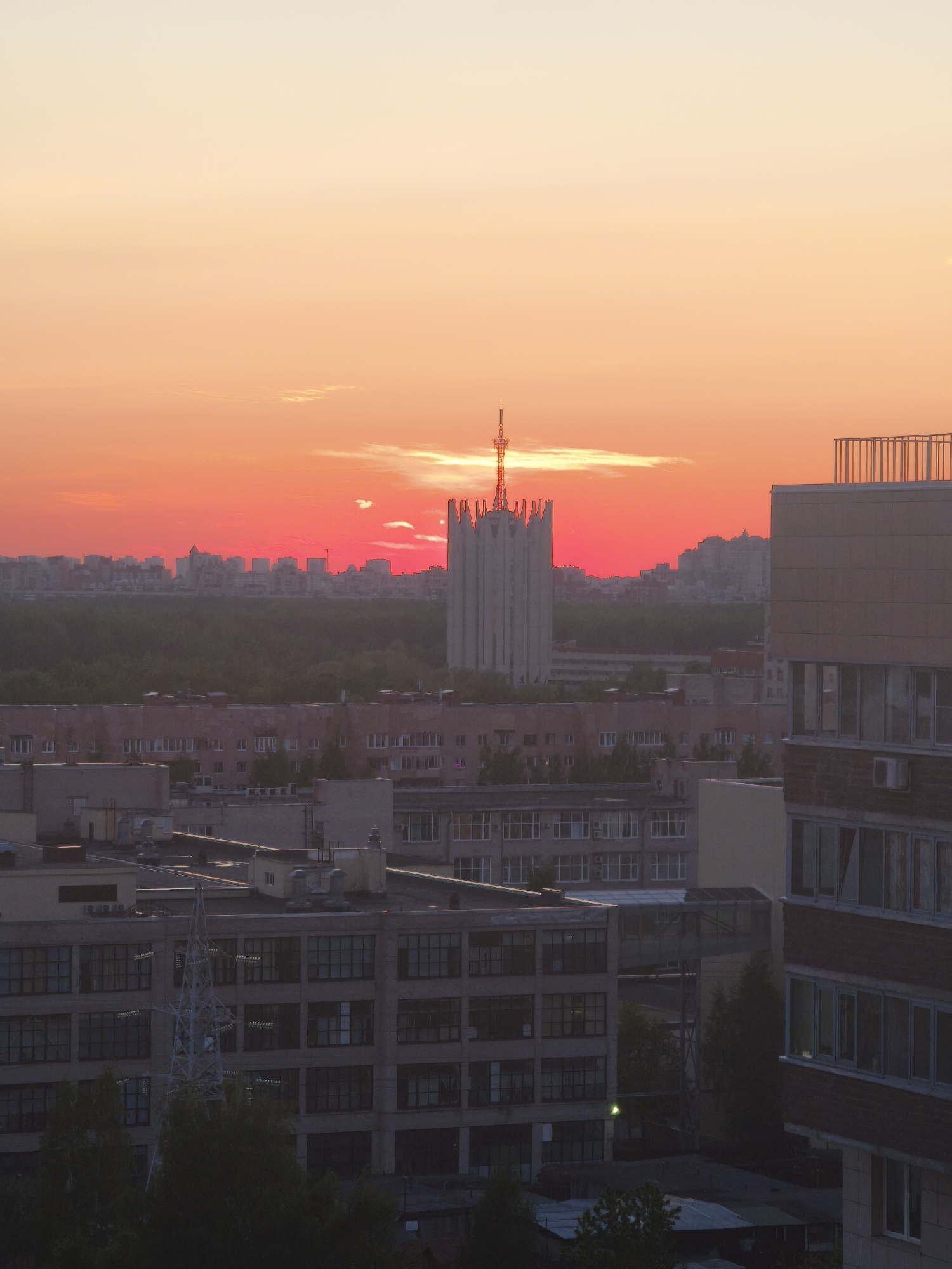 The image captures a cityscape at sunset in Saint Petersburg, Russia. The sky transitions from a bright orange near the horizon to a pale yellow at the top. The primary focal point is a tall, light-colored building in the mid-ground with a distinctive spire atop, resembling a broadcasting or communications tower. Below this building, there is a line of trees, likely a park or wooded area, that creates a visible separation.
In the foreground, several low-rise, multi-story buildings are visible, typical of urban or industrial architecture. These buildings are mostly gray and beige. There are trees in the foreground, along with an electrical transmission tower. A tall residential building is visible in the right of the shot.