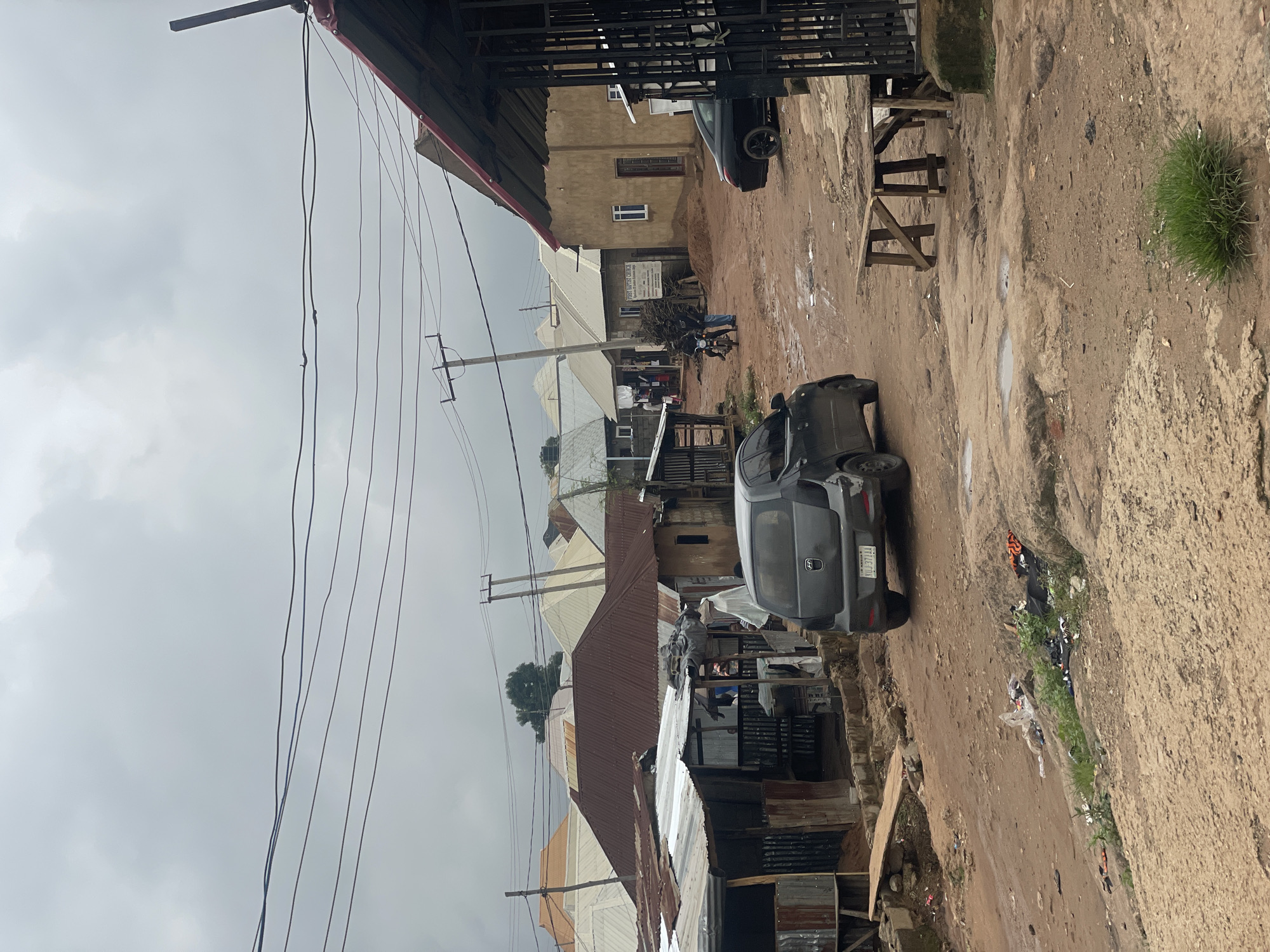 This is a medium shot, eye-level, outdoor, daylight photograph of a street scene in Kurunduma, Nigeria. The sky is overcast and gray. In the foreground, the street is made of dirt with some puddles of water. To the right, there is a wooden table with two chairs. Further back, a dark gray hatchback car with license plate "KUJ 371AA" is parked on the right side of the street. To the left, there are several low-rise buildings with corrugated metal roofs, some of which are rusty. Some of these buildings appear to be shops or homes. In the background, more buildings with various roof types are visible, along with utility poles and overhead wires. Several people are present, including a person wearing a white garment and a headscarf walking near the shops. A motorcycle with two individuals on it is also visible on the street. The overall environment appears somewhat dilapidated, with some scattered debris and untended vegetation.