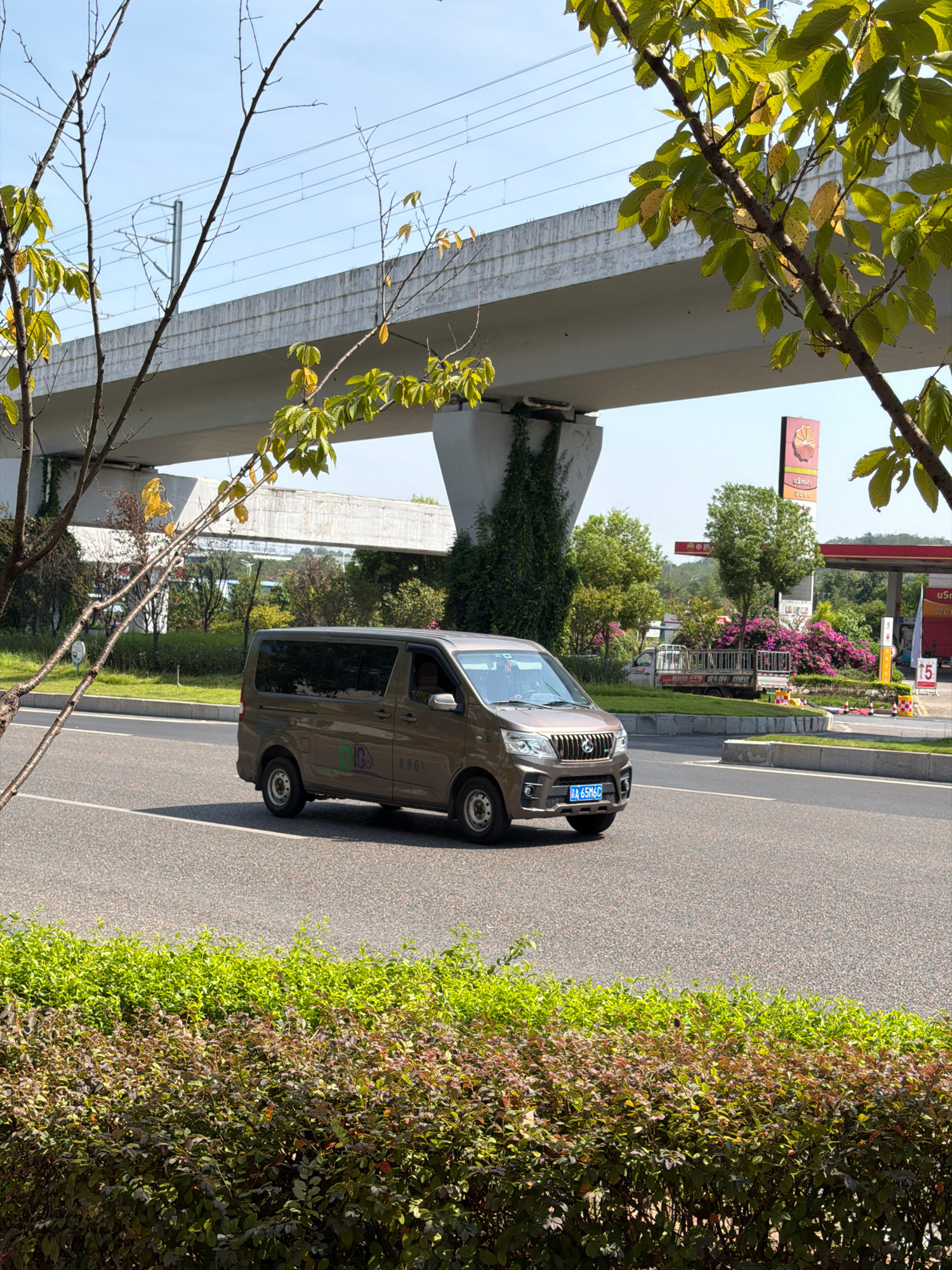 In Jiangjin, China, a brown minivan with a license plate reading "A 65M6C" drives along a paved road beneath an elevated concrete structure. The minivan has tinted windows and some graphics on its side, including a green circle with a white symbol. The road is bordered by green bushes in the foreground and trees and flowering bushes in the background. A gas station with a red canopy and signs is visible to the right, along with a small utility truck. Overhead power lines are present, and the sky is clear and blue.