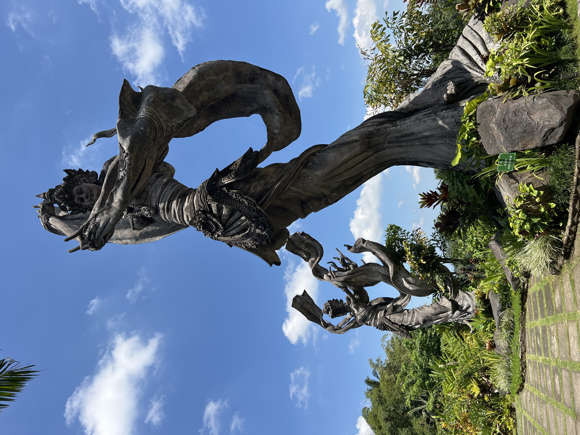 The image depicts a monumental sculpture in Kedewatan, Indonesia. Two figures are sculpted in grey material against a background of a clear blue sky with scattered white clouds. The upper figure is heavily adorned with ornamentation, including a headdress and intricate details on the body. It is dramatically posed as if in motion or flight, with flowing fabric wrapping around it. The lower figure echoes the style and posture of the upper one, though partially obscured by the angle. Lush greenery and garden landscaping surround the base of the sculpture, with paving stones indicating a walkway. The sculpture is situated outdoors in a public area.