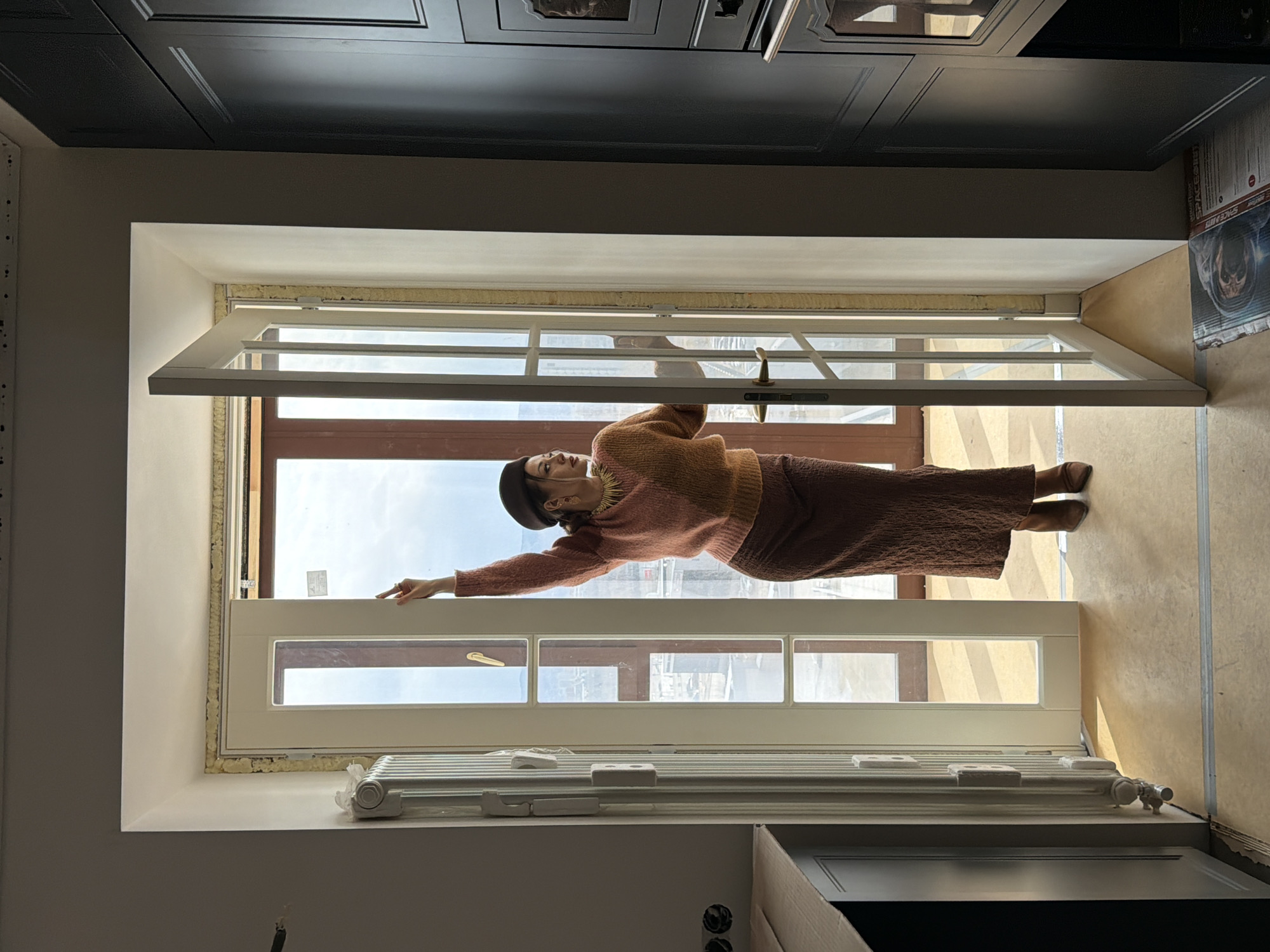 A woman is standing in a window frame in an interior setting. She is wearing a brown beret, an orange and brown sweater, and a brown skirt. The window frame is white and the windows are open, revealing a view of what appears to be an urban landscape with buildings and sky. A white radiator is visible below the window. Above the window is a dark cabinet. The interior appears to be undergoing renovation. I cannot ascertain the location is in Krasnoyarsk, Russia.