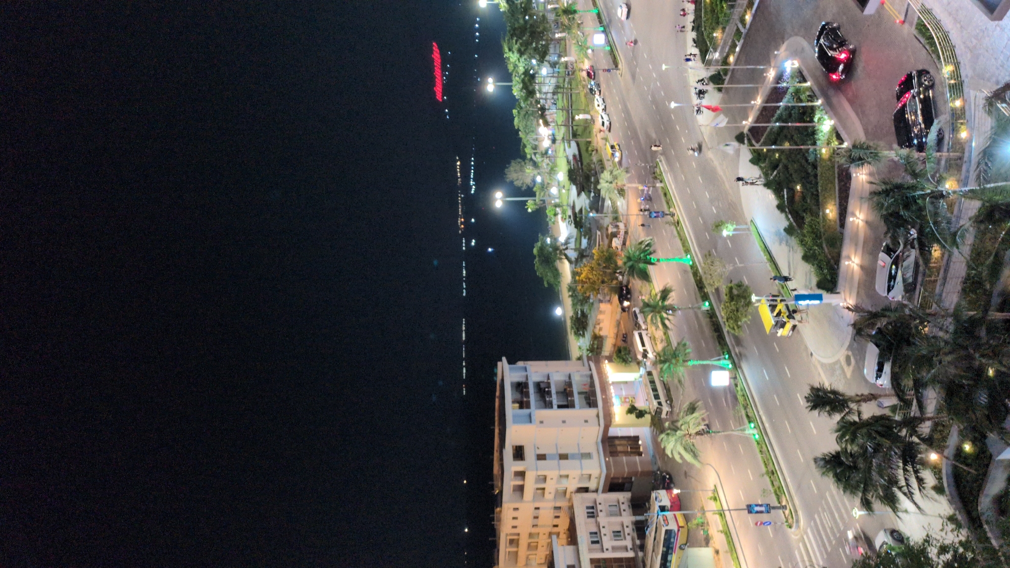 The image depicts a nighttime scene of a coastal area in Quy Nhon Ward, Vietnam, viewed from an elevated perspective. The foreground features a landscaped area with palm trees and a circular driveway where multiple vehicles, including black, white, and red passenger cars, are present. Several flags on poles are erected within this area.
Immediately beyond, a multi-lane asphalt road runs parallel to the coastline. It is divided by a central median adorned with palm trees, some of which are illuminated with green light. Various vehicles, including cars, white vans, a yellow and red bus on the far left, and a distinct yellow open-sided vehicle stopped near a crosswalk, are visible on the road. Pedestrians are walking on the sidewalks and near the yellow vehicle. Streetlights illuminate the roadway and sidewalks.
To the left of the road, a multi-story building with multiple balconies stands prominent, exhibiting illuminated windows. Further along the coastline, a landscaped beachfront park area is visible with additional palm trees and streetlights.
The background consists of a vast, dark body of water, likely the ocean or a bay, extending to the horizon. Numerous small, scattered lights are visible across the water, possibly from distant vessels or land. A large red neon sign, partially legible as "Anantara" or similar, is faintly visible on the distant horizon above the water. The sky above is dark, indicative of nighttime.
