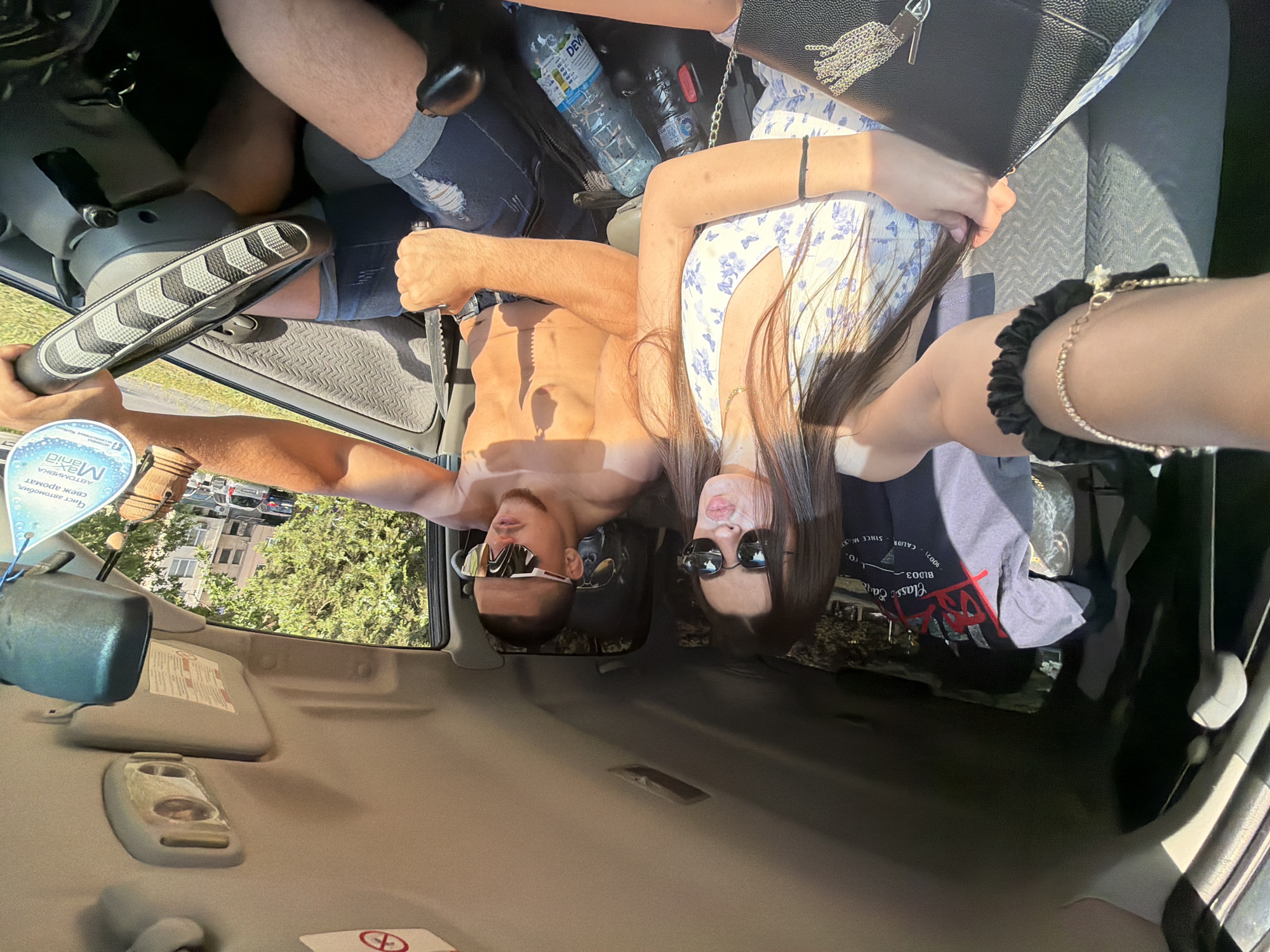 An interior shot of a vehicle shows a female in the front passenger seat taking a selfie, and a male in the driver's seat. The scene is illuminated by strong daylight, indicating it is daytime in an urban environment in Plovdiv, Bulgaria.
The female, positioned on the right side of the image, has long dark hair and wears dark, round-framed sunglasses. She is dressed in a white halter-neck top with a light blue floral (butterfly) pattern. Her right wrist features a black scrunchie and a gold chain bracelet with a charm. A gold chain necklace is also visible. She holds a black purse with a gold chain tassel in her left hand, which also clutches strands of her hair. Her lips are pursed. Her right arm is extended, holding the camera for the selfie.
The male, in the driver's seat, has short dark hair and stubble, wearing light-colored, athletic-style sunglasses. He appears shirtless, showcasing a tanned physique, and wears ripped denim shorts with a black belt. His left hand firmly grips a fixed-blade knife with a serrated edge and a black handle. His right hand rests on the steering wheel, which is fitted with a black and gray textured cover. He is looking forward, not at the camera.
The car's interior features a light-colored headliner with an overhead light fixture and sun visors; a "no smoking" symbol is visible on the passenger's sun visor. The seats are upholstered in a gray fabric with a subtle chevron pattern. Between the front seats, at least two plastic water bottles, one branded "DEWA", are visible. A black gear shift is located to the male driver's right. Draped over the back of the driver's seat is a gray t-shirt with red and white text, including "Classic Earth," "BLD03," and "CALIF. SINCE M.L." A blue-tinted rearview mirror is present, from which hangs a circular blue air freshener with Cyrillic text reading "Чист автомобил, свеж аромат" (Clean car, fresh aroma) and "автомивка Max..." (car wash Max...). Through the car windows, trees, residential apartment buildings, and other parked vehicles are discernible, characteristic of an urban setting.