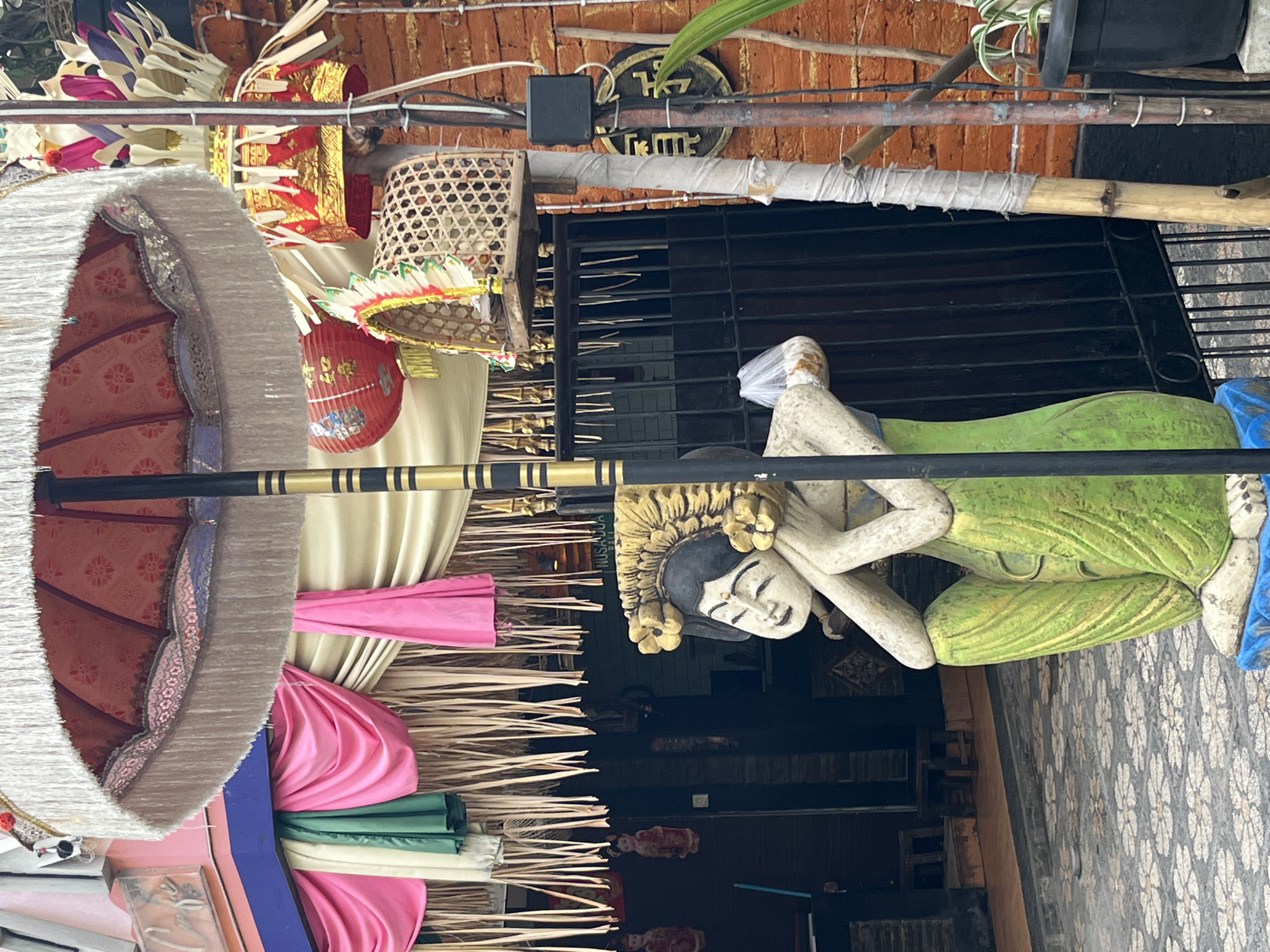 A statue of a seated woman with a serene expression, her head resting on her hands, is positioned in front of a black metal gate and a brick wall. The statue wears a gold headdress adorned with flowers and a green garment. Behind the statue, a large decorative umbrella with white fringes is open, its patterned fabric visible. Numerous colorful fabrics are draped above the entrance to a building, creating a festive atmosphere. Red lanterns and decorative offerings are also visible, suggesting a cultural or religious event. The ground is paved with tiles featuring a floral pattern. The scene is located in Kuta, Indonesia, a popular tourist destination known for its beaches and vibrant culture.
