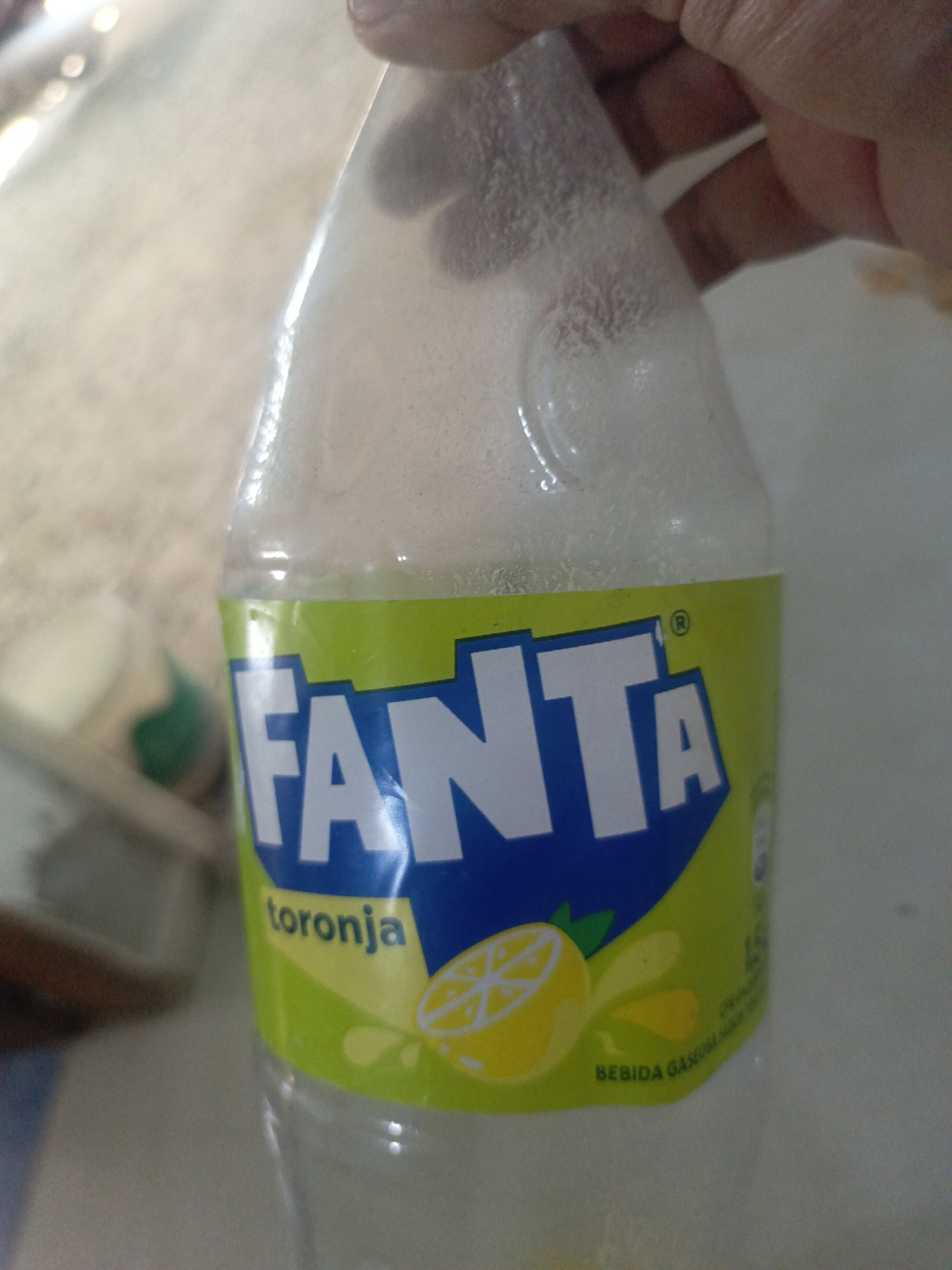 A close-up shot shows a partially empty bottle of Fanta Toronja (grapefruit flavor) soda, likely in Juan Griego, Venezuela, given the Spanish labeling. The bottle is being held, and the top portion appears frosted or contains condensation. The Fanta logo is prominent, with a slice of grapefruit depicted on the label.