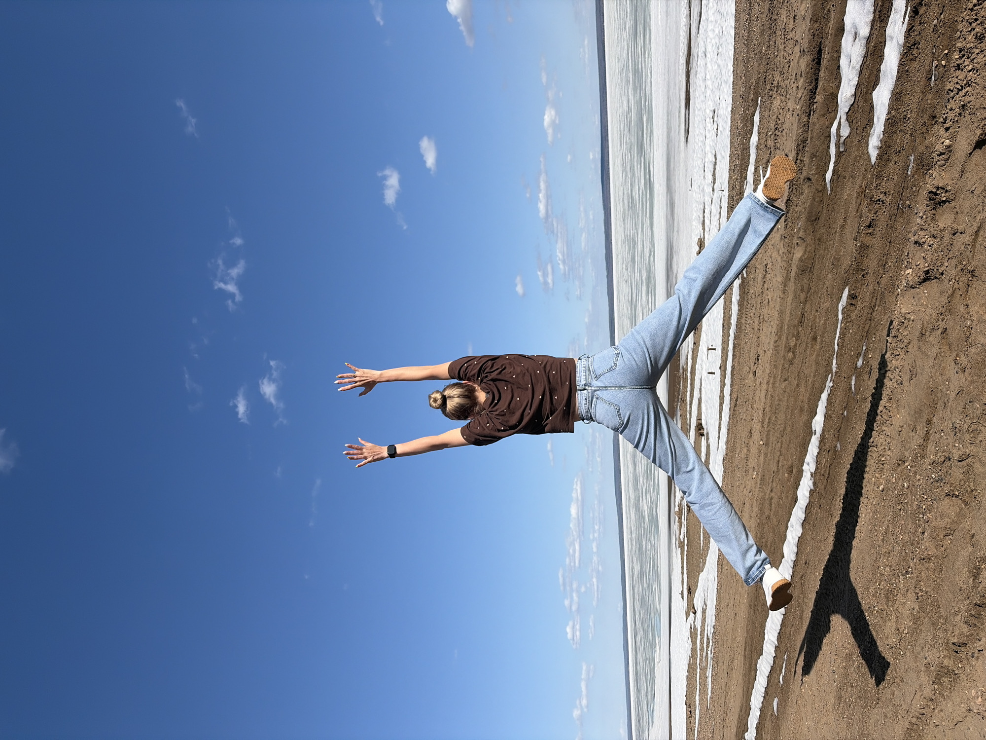 A woman stands on a brown dirt bank, extending her arms and legs outward to form a 'Y' shape against a blue sky with scattered clouds. She wears a dark brown shirt with white polka dots, light blue jeans, and white shoes with brown soles. Her hair is pulled back in a bun. A frozen lake or river bordered with patches of snow is visible. The location is outdoors in Noviy, Russia.