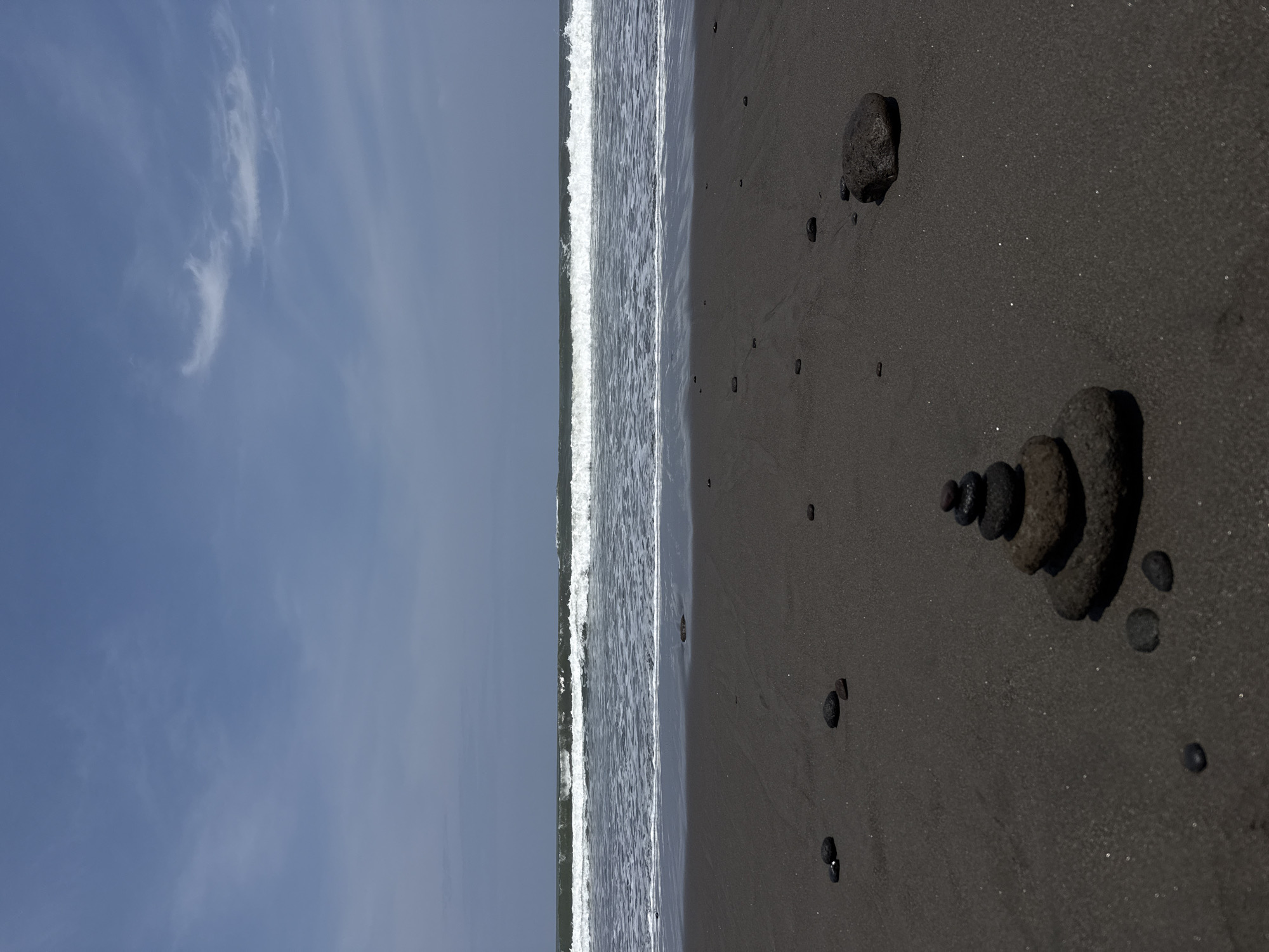 This image captures a coastal scene in Sudimara, Indonesia. The lower third of the frame consists of a dark grey, volcanic sand beach, appearing damp and exhibiting a subtle sheen. Centered in the lower foreground is a cairn composed of five dark, rounded stones stacked vertically, with the largest stone forming the base. Numerous other dark, rounded stones are scattered across the wet sand, particularly around the cairn and extending to the mid-right where a larger, isolated dark stone rests. The midground features the ocean, with a prominent line of white breaking waves stretching horizontally across the frame, receding into darker grey-blue water towards the horizon. The upper third of the image displays a light blue sky with scattered, wispy white cloud formations. No human subjects, animals, or man-made structures are visible.