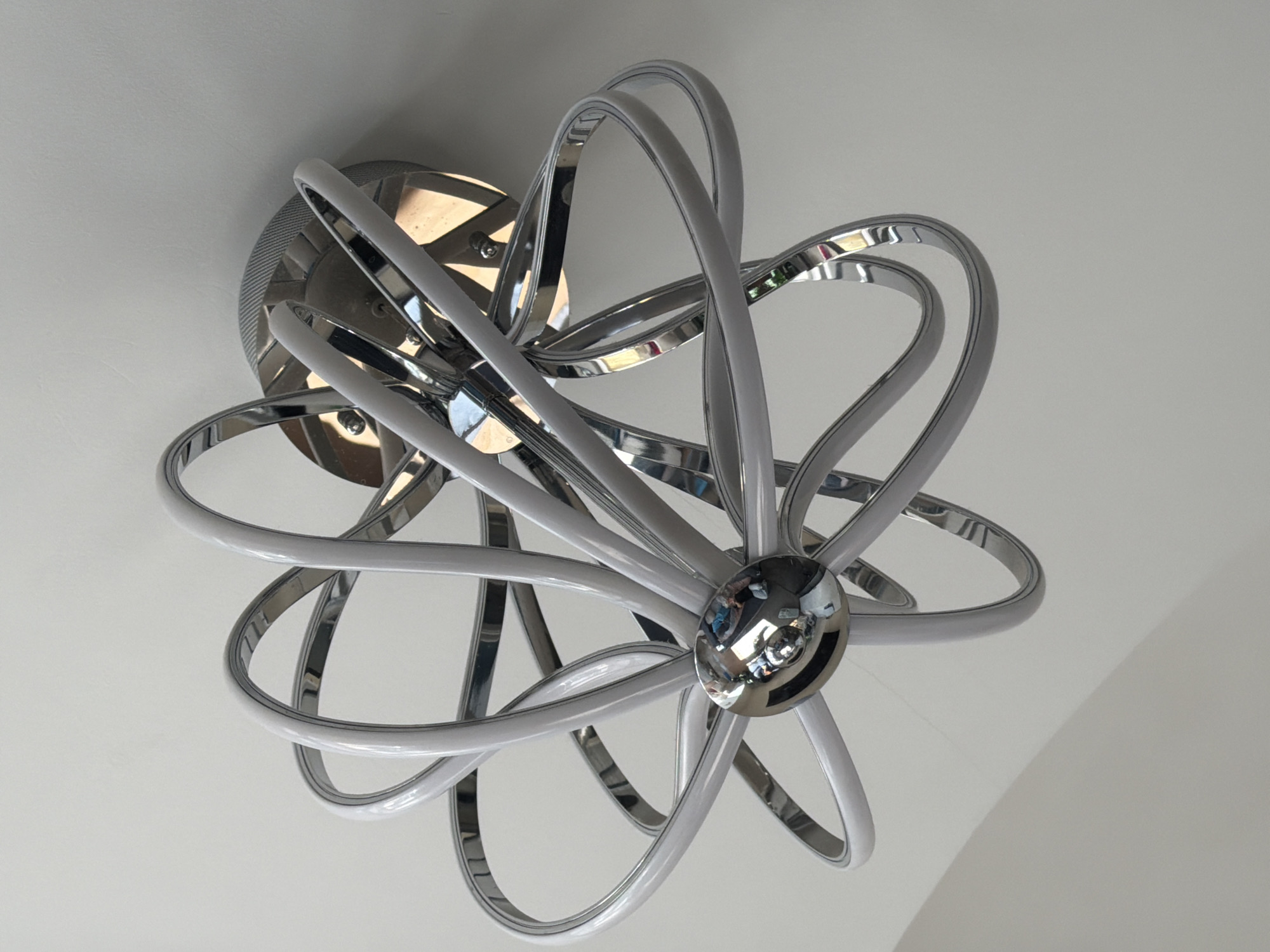 Here is a description of the image:
The image is a close-up, upward-facing view of a modern chandelier attached to a white ceiling. The chandelier features a central chrome-colored sphere and multiple intersecting, looping arms. Each arm is constructed with two parallel chrome strips encasing a white light strip. The light fixture is mounted to a chrome-colored base. The surrounding ceiling is plain white. The location is Unknown, Unknown.