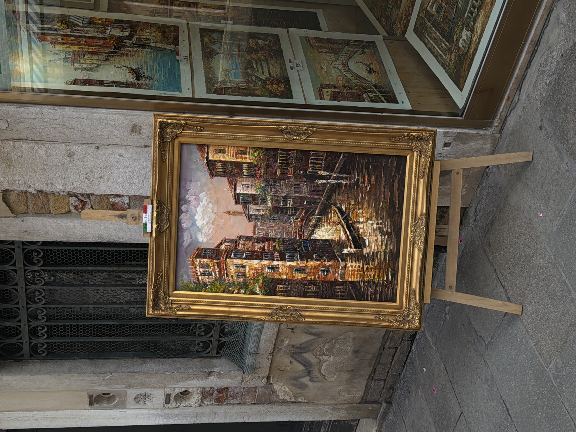 A framed painting of a Venetian canal scene is displayed on a wooden easel on a cobbled street in Venice, Italy. The painting, rendered in a textured, oil-on-canvas style, depicts tall, colorful buildings lining a narrow waterway. A gondola glides through the water, reflecting the warm hues of the sunset sky. The easel is positioned in front of a building with a wrought-iron-grated window and textured stone facade. To the right of the easel, another building with a large display window shows several more paintings, some reflecting the street scene. The ground is paved with stone, and a few pink flower petals are scattered on the pavement. The overall atmosphere is that of an outdoor art display in a historic Italian city.