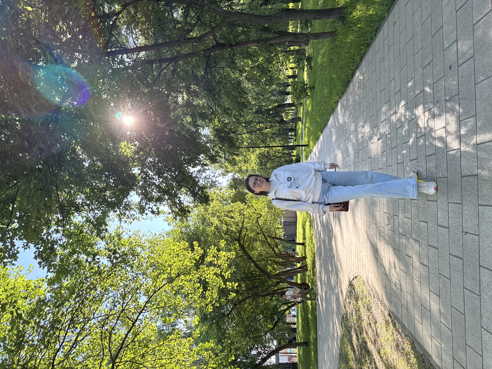 An eye-level, medium shot captures a young woman standing on a paved walkway in a park. She is wearing a white hoodie, light blue jeans, and white sneakers. The woman is smiling and looking at the camera. Behind her, the walkway extends into the distance, lined with green grass and trees. Sunlight streams through the leaves of the trees, creating dappled shadows on the walkway. In the background, another person is walking away from the camera on the same walkway. The scene appears to be in a city park, possibly in Izhevsk, Russia, given the context. The overall atmosphere is bright and cheerful, suggesting a pleasant day outdoors.