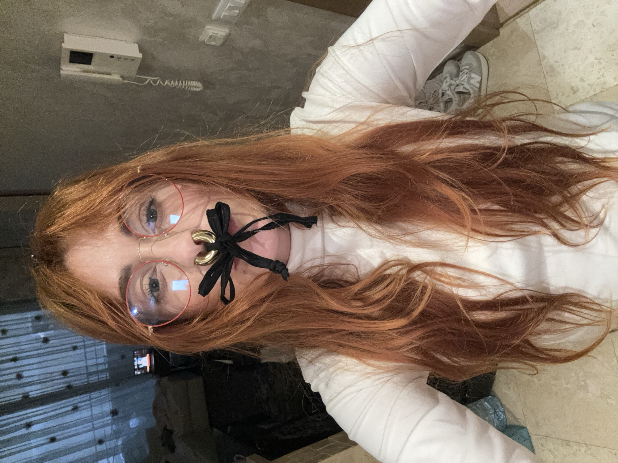 An indoor, close-up, chest-up selfie depicts a female in Budva, Montenegro, with long, reddish-brown hair, wearing red-rimmed glasses and a white long-sleeved top. She has a large, golden, circular septum piercing from which a black ribbon is tied in a bow, covering her mouth. Her light-colored eyes are directed at the camera. The background to the left shows dark, vertical window coverings and a small screen displaying an image. To the right, a textured grey wall features a white wall-mounted intercom system with a coiled cord and a white electrical outlet below it. On the light-colored tiled floor, a pair of grey New Balance sneakers is partially visible.