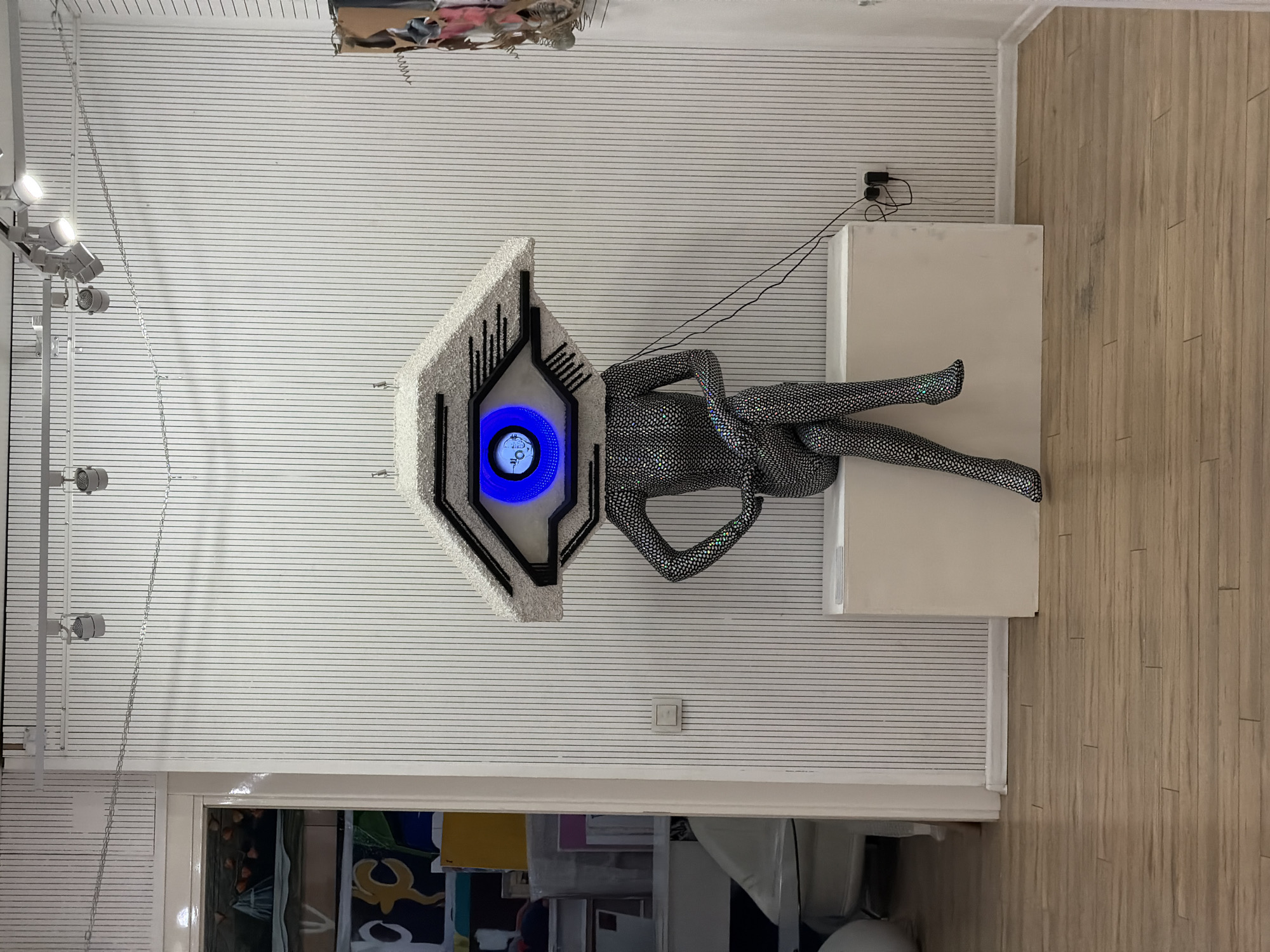 This image depicts an indoor display featuring a futuristic, mannequin-like figure in a modern setting. The main subject is a reclining figure positioned on a white rectangular pedestal.
The figure possesses a distinctive, angular head, predominantly white with black accents, reminiscent of a sleek, abstract helmet or robot head. Centrally placed on its head is a bright, circular blue light, acting as an eye or display screen. Within this blue circle, white digital text is visible: "4:00" on the upper line, and below it, "BY" (or potentially "BV") followed by "°C", suggesting a time and temperature readout. The body of the figure is covered in a shimmering, textured material, possibly speckled silver and black fabric, giving it a high-tech or alien appearance.
The room's wall has a subtle horizontal striped pattern. On the left side of the wall, a track lighting system with multiple spotlights illuminates the display. Behind the white pedestal, a black power adapter is mounted on the wall, with black wires extending from it. A simple white light switch is also visible on the wall. The floor to the right features light-colored wooden planks. At the bottom of the image, a doorway leads to an adjacent room, revealing indistinct shapes and colors, possibly furniture or other items. A piece of abstract art or a cardboard box structure is partially visible in the upper right corner of the wall. The scene is static, showcasing an artistic or commercial installation, with no active event taking place.